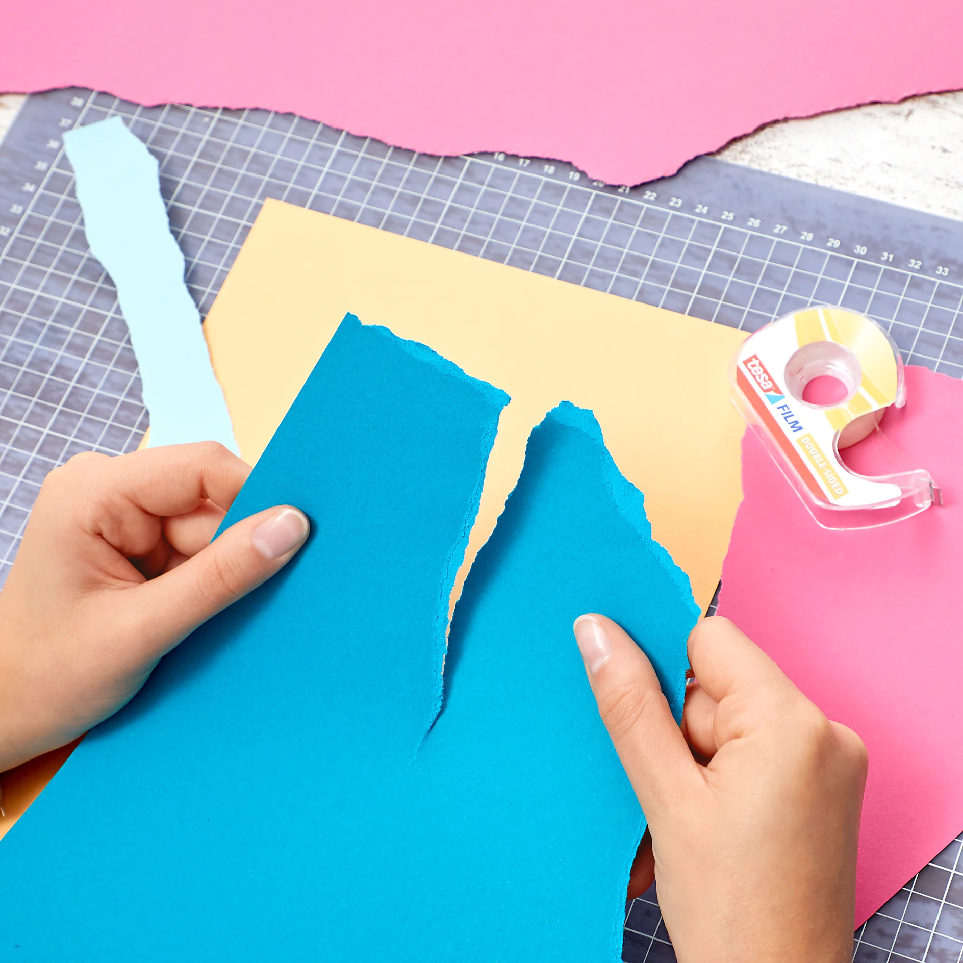 Hands are tearing a blue sheet of paper over a grid-lined cutting mat. Nearby are other torn paper sheets in pastel colors: yellow and pink. A roll of clear tesa tape in a plastic dispenser is also in view. (This text has been generated by AI)