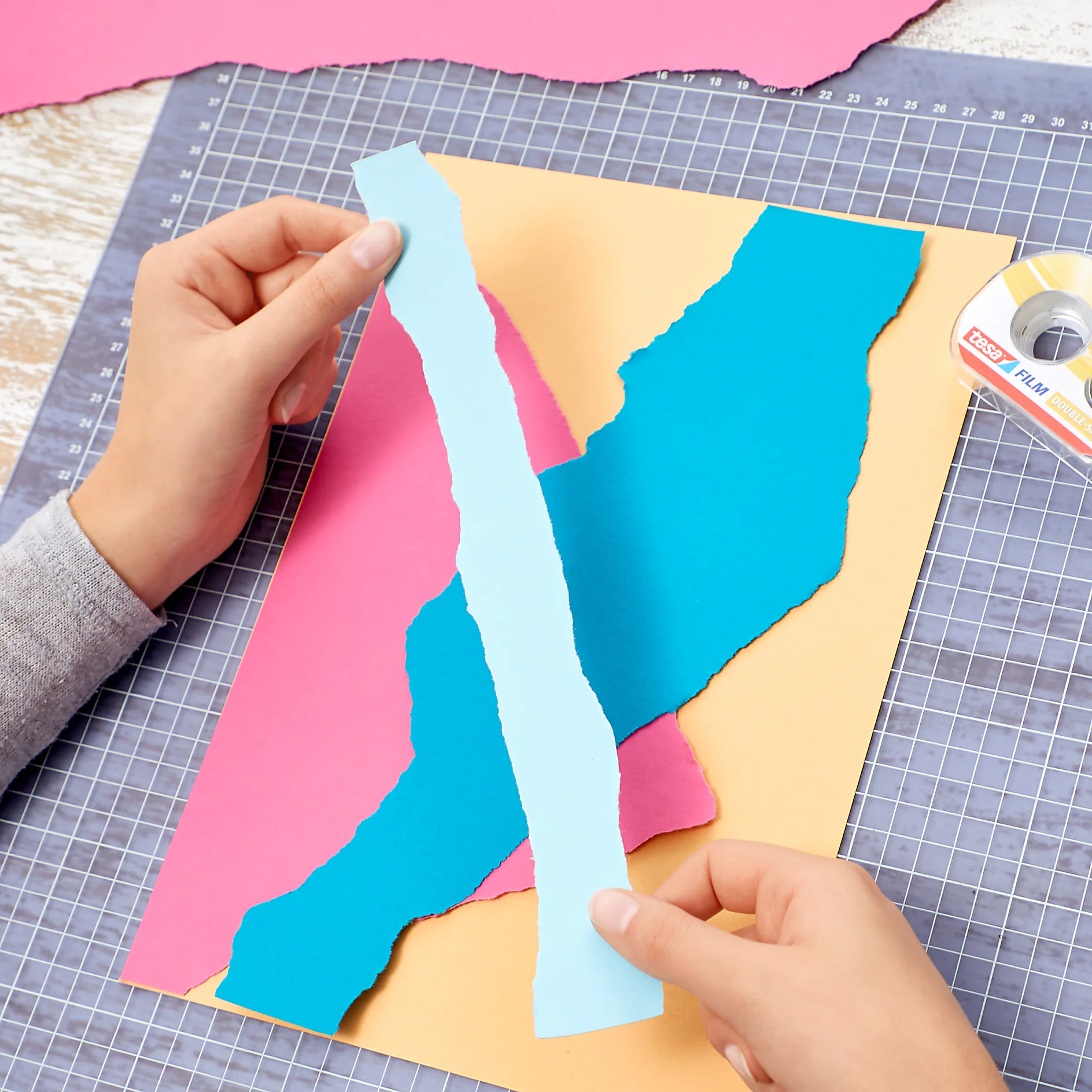 Hands are arranging torn strips of pink, blue, and teal paper on a yellow sheet. The background features a cutting mat. A roll of tesa tape is visible to the side. (This text has been generated by AI)