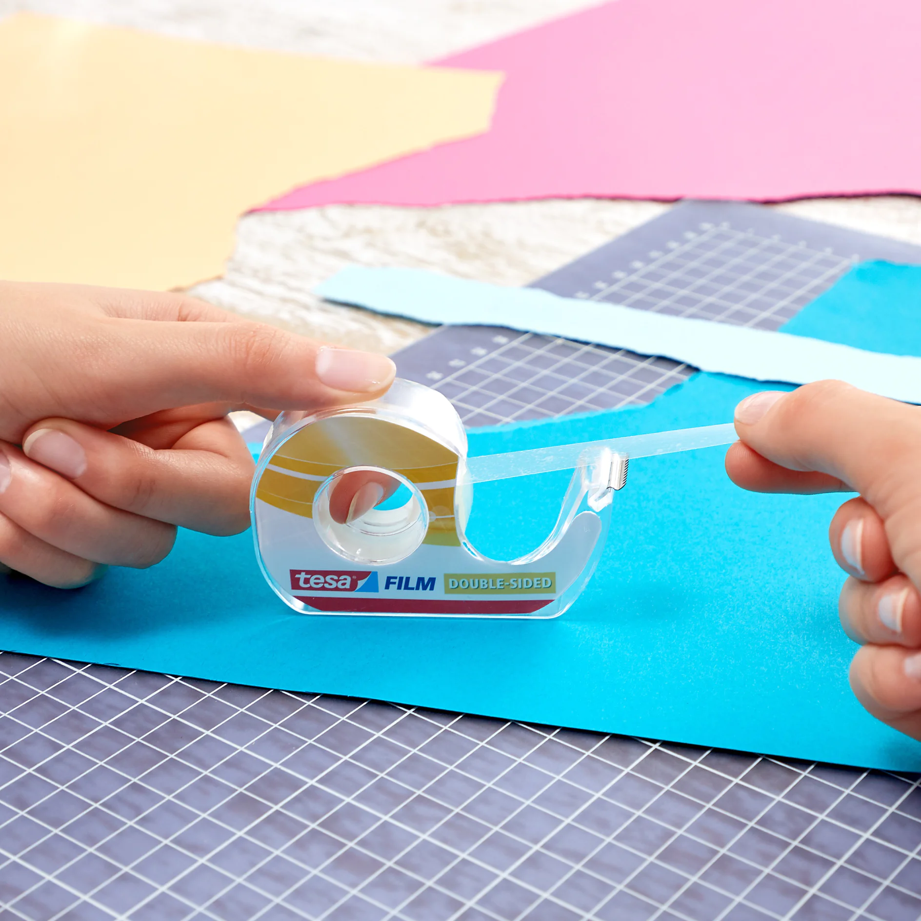 Two hands using a roll of double-sided tesa tape labeled tesa FILM on a craft project. The tesa tape is being pulled from the dispenser onto a sheet of blue paper, with pink and yellow papers nearby on a grid-patterned surface. (This text has been generated by AI)