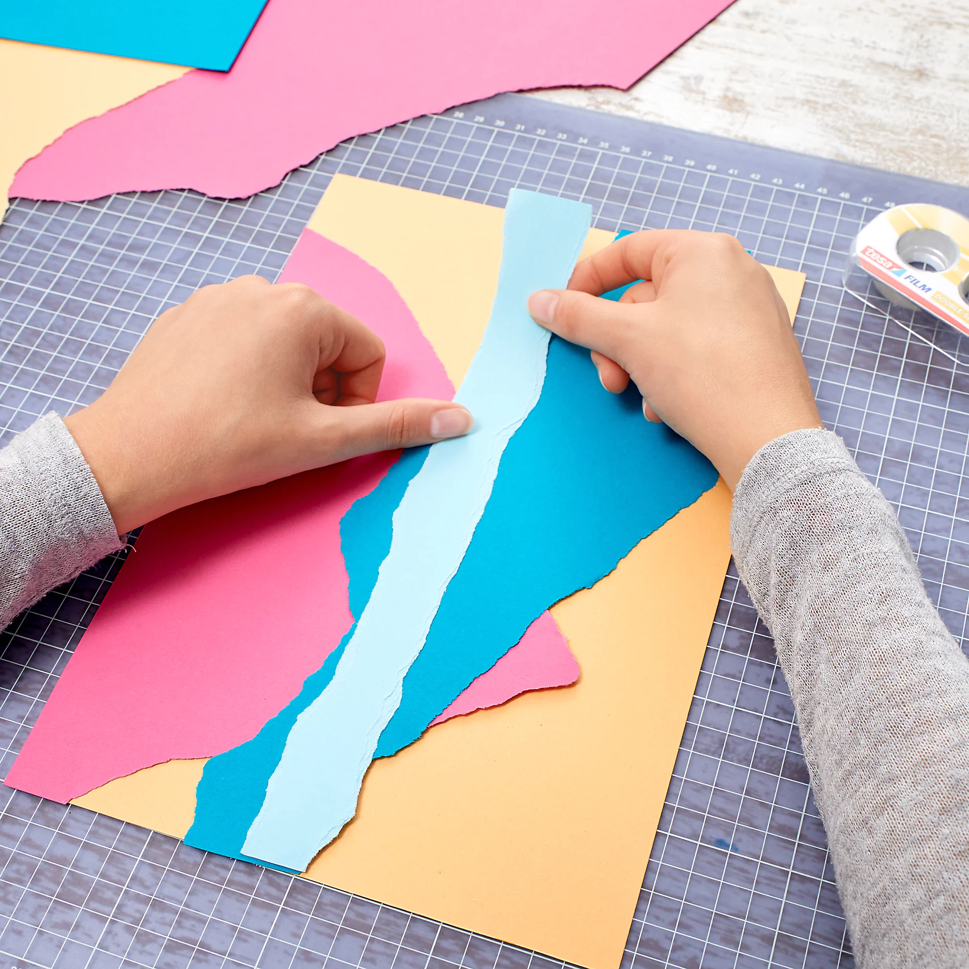 Two hands arranging torn pieces of colored paper (pink, blue, yellow) on a grid cutting mat. A roll of tesa tape is in the upper right corner. (This text has been generated by AI)