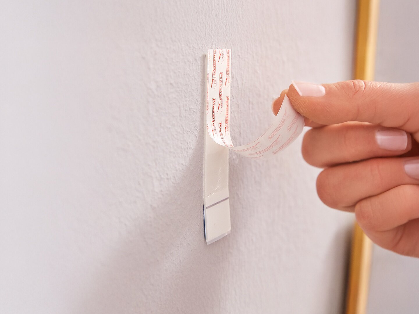 Una mano está despegando el adhesivo de un gancho de pared de plástico blanco que está fijado a una pared de color claro usando cinta tesa. (This text has been generated by AI)
