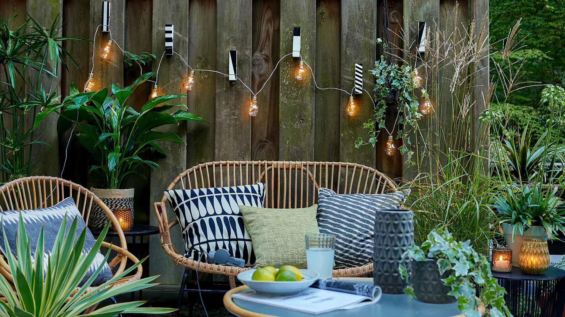Una acogedora zona de descanso al aire libre cuenta con sillas de mimbre con cojines estampados, una mesa decorada con velas y limas, todo rodeado de diversas plantas verdes. Luces de cadena cuelgan en la valla de madera del fondo, sujetas con cinta tesa, creando un ambiente cálido y acogedor. (This text has been generated by AI)