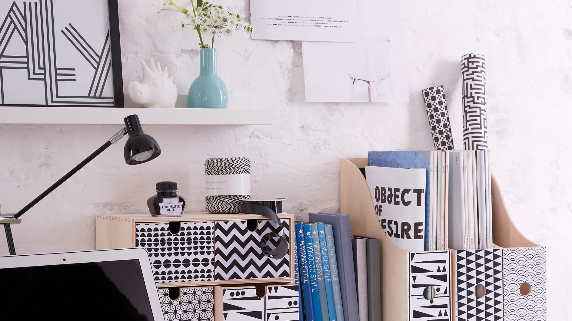 Configuración minimalista de escritorio con un portátil, cajas estampadas y una lámpara sobre una superficie blanca. Los libros y objetos decorativos están organizados ordenadamente. El fondo es una pared de ladrillo blanco con pósteres y una pequeña estantería que exhibe un jarrón y arte enmarcado, todo perfectamente sujeto con cinta tesa. (This text has been generated by AI)