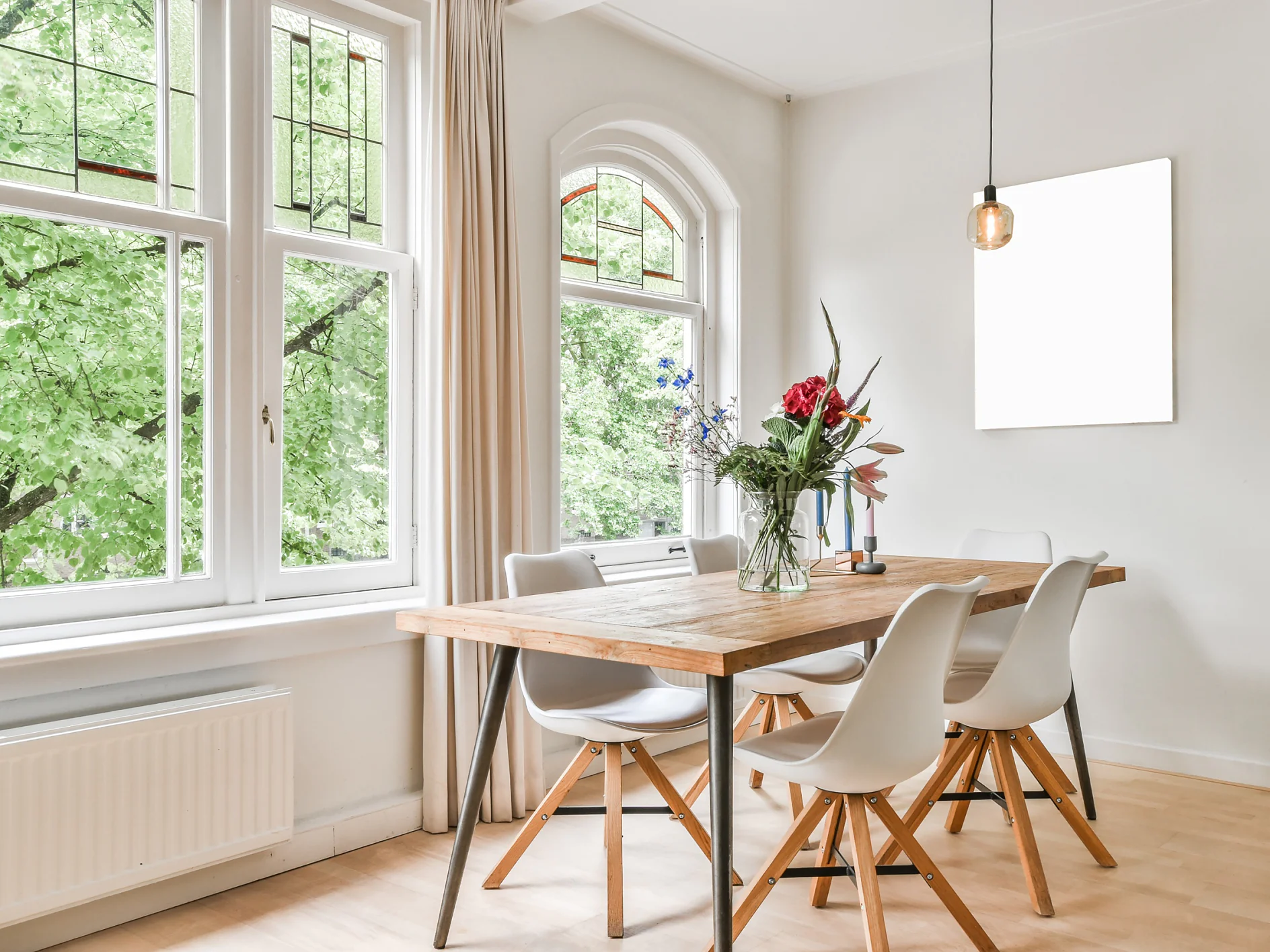 Un comedor luminoso cuenta con una mesa de madera acompañada por seis sillas blancas. Sobre la mesa hay un jarrón lleno de flores. A la izquierda, grandes ventanales ofrecen vistas a árboles verdes. La decoración incluye una sola lámpara colgante y un marco vacío en la pared. El suelo es de madera. (This text has been generated by AI)