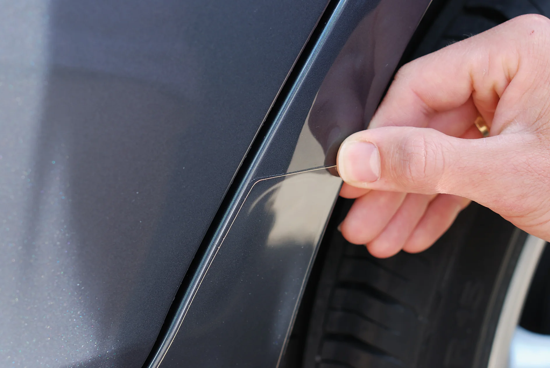 mano despegando una lámina protectora transparente del guardabarros de un coche gris oscuro cerca del neumático en un entorno exterior (This text has been generated by AI)
