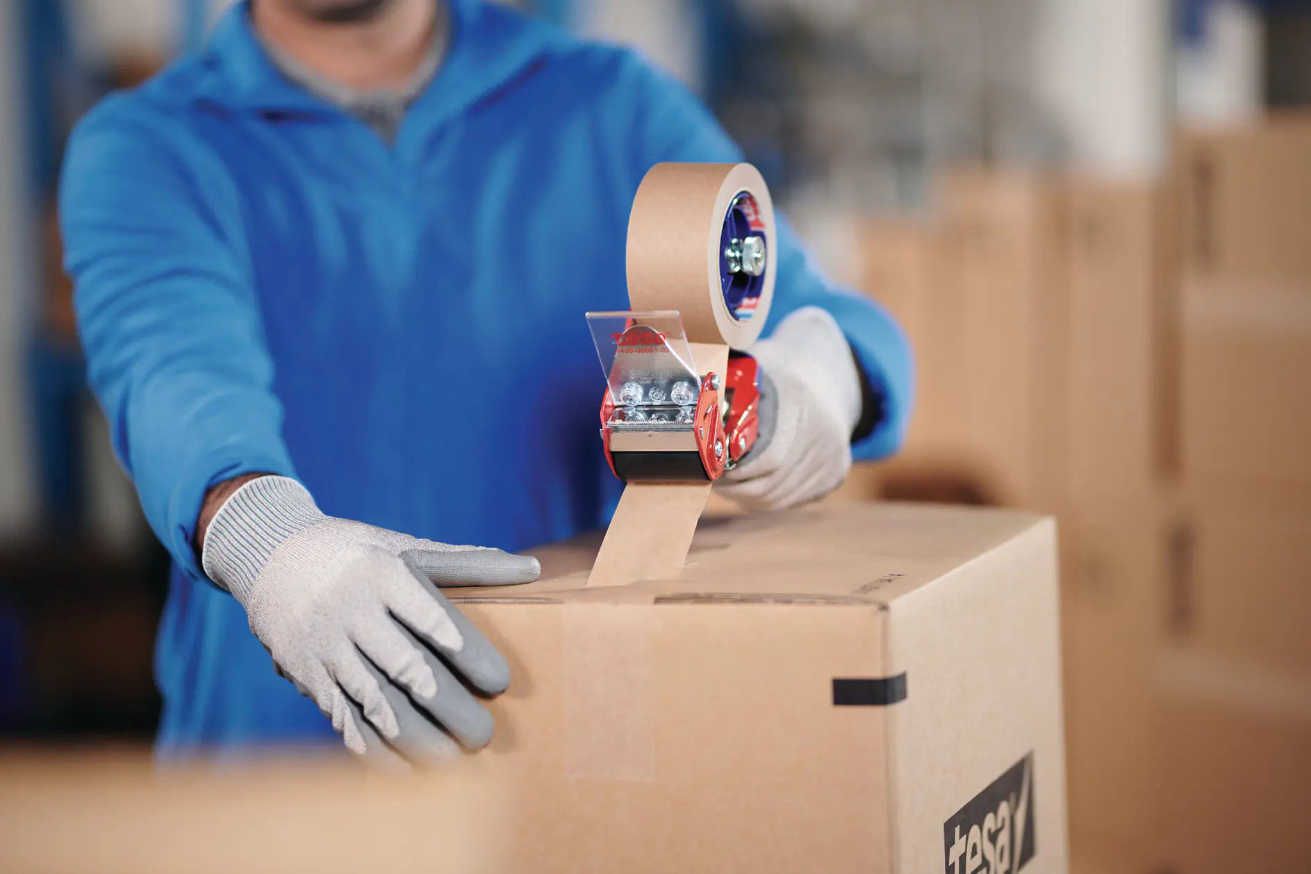 person wearing gloves sealing a cardboard box with tesa tape in a warehouse setting (This text has been generated by AI)