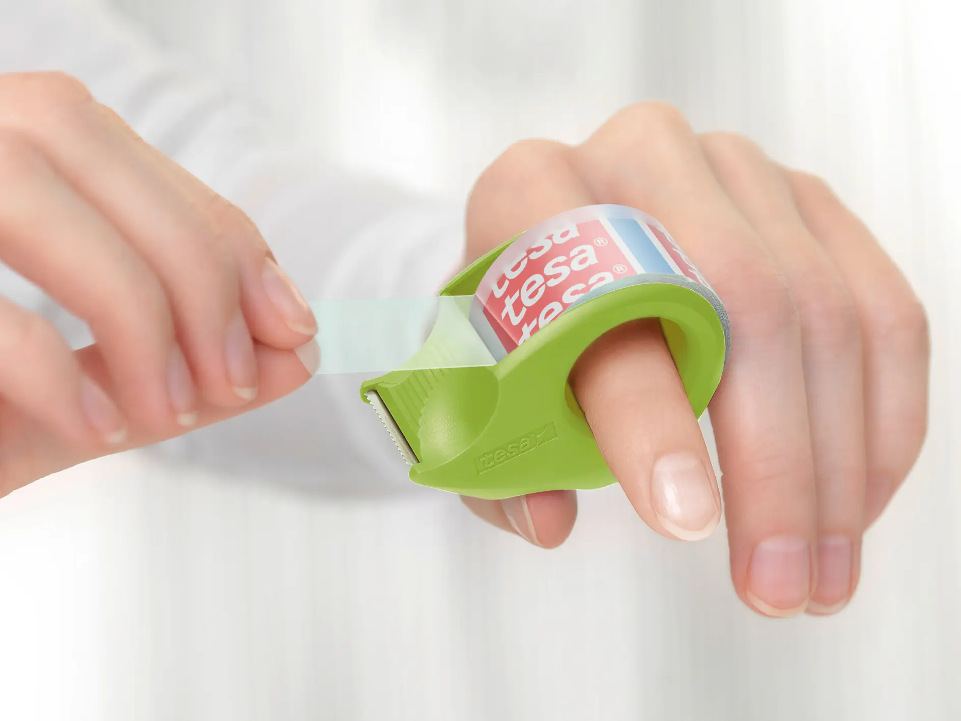 Hands using a green tesa tape dispenser to pull clear adhesive tape, set against a blurred white background. (This text has been generated by AI)