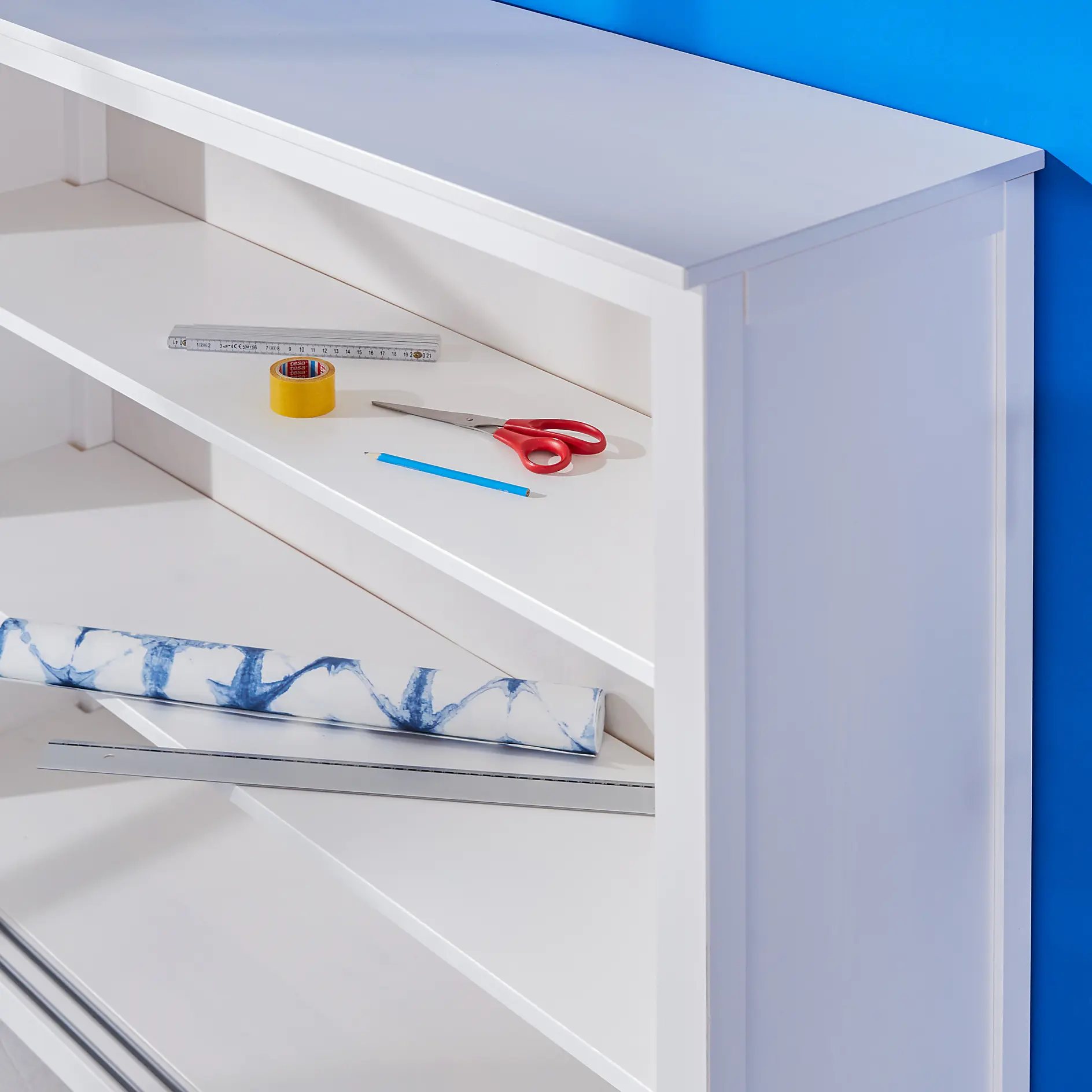 A white bookshelf against a blue wall with items on the top shelf, including a metal ruler, yellow tesa tape with a red center, red scissors, and a blue colored pencil. A folded, blue and white fabric or paper is on a lower shelf. (This text has been generated by AI)