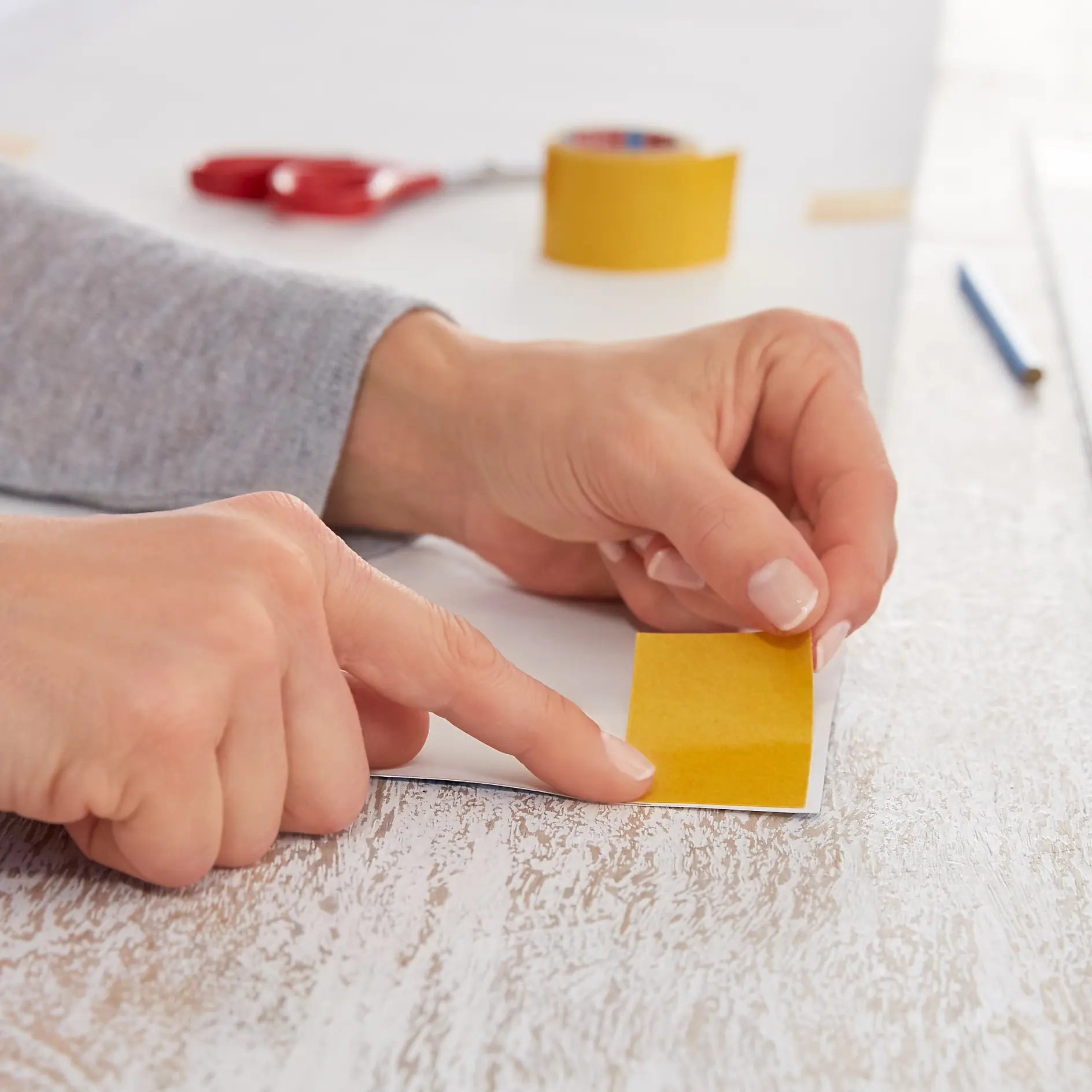 Close-up of hands peeling the backing off a piece of yellow tesa tape on a white surface. A roll of tesa tape, red scissors, and a pencil are on the wooden table in the background. (This text has been generated by AI)