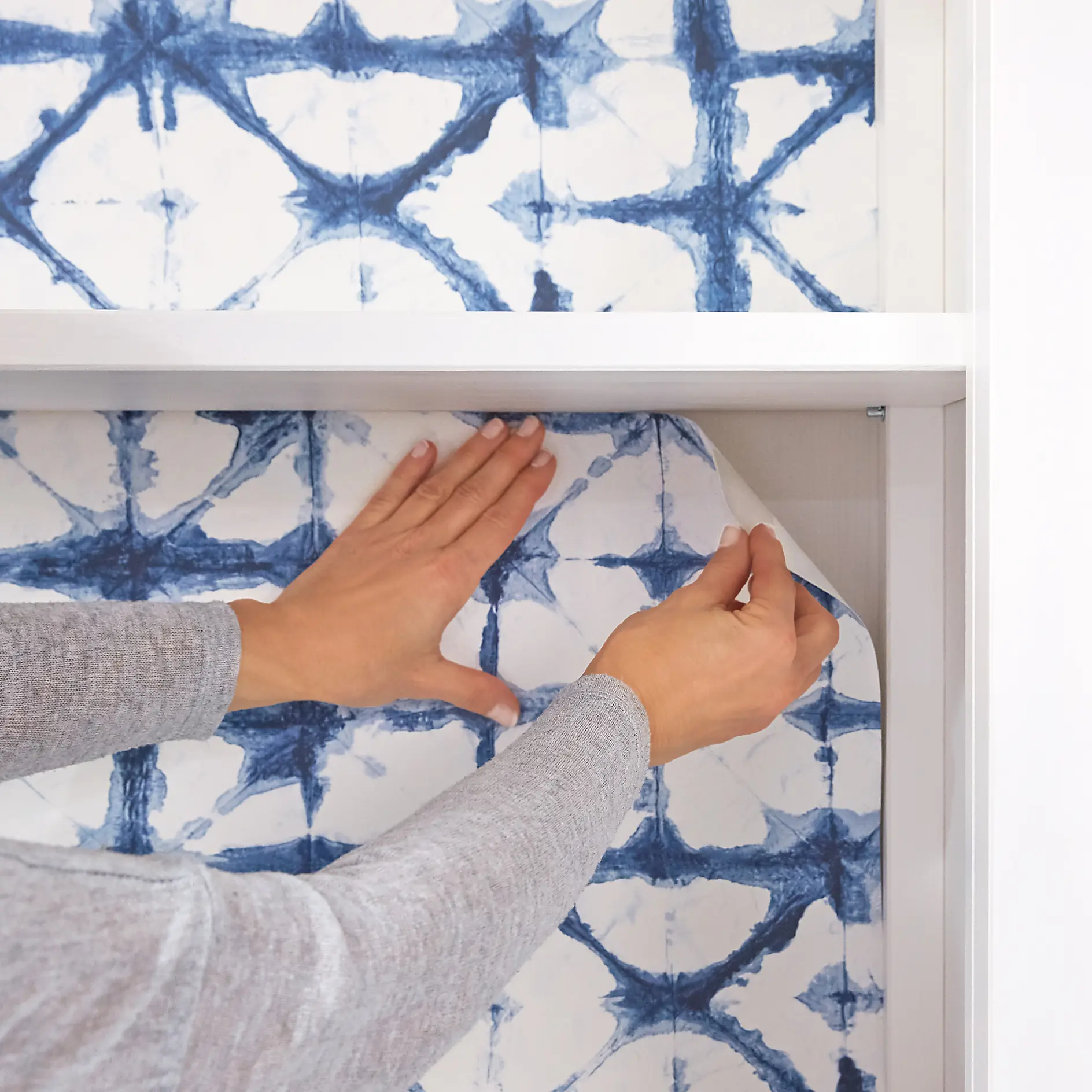 Two hands peel off blue and white patterned adhesive wallpaper from a wall. The pattern features dark blue intersecting lines on a white background. The persons sleeves are light gray, and the background includes a white, smooth surface. To assist in the process, they use tesa tape for additional precision and ease of removal. (This text has been generated by AI)