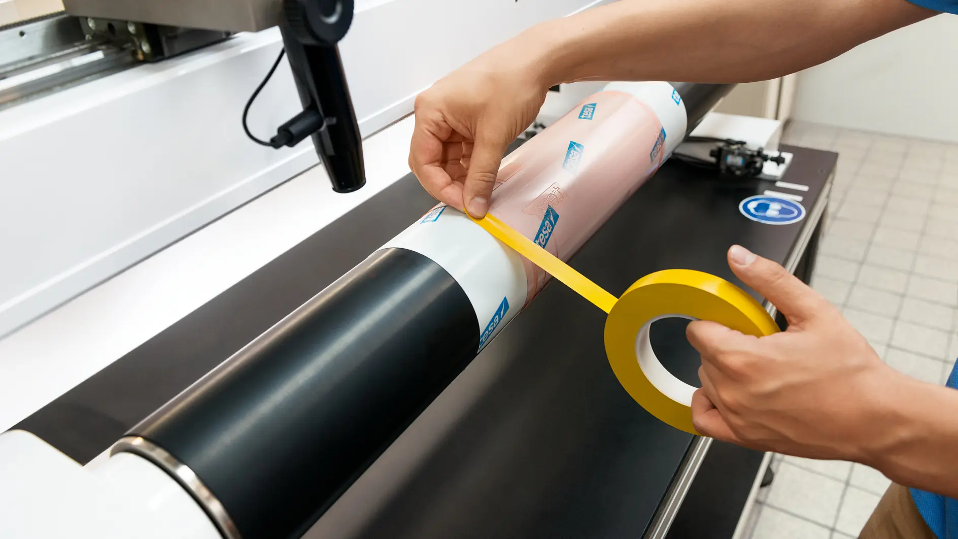 Person applying yellow tesa tape to a cylindrical object on a machine, surrounded by a workspace with a tiled floor. The object includes two sections, one black and one pink with blue logos. (This text has been generated by AI)