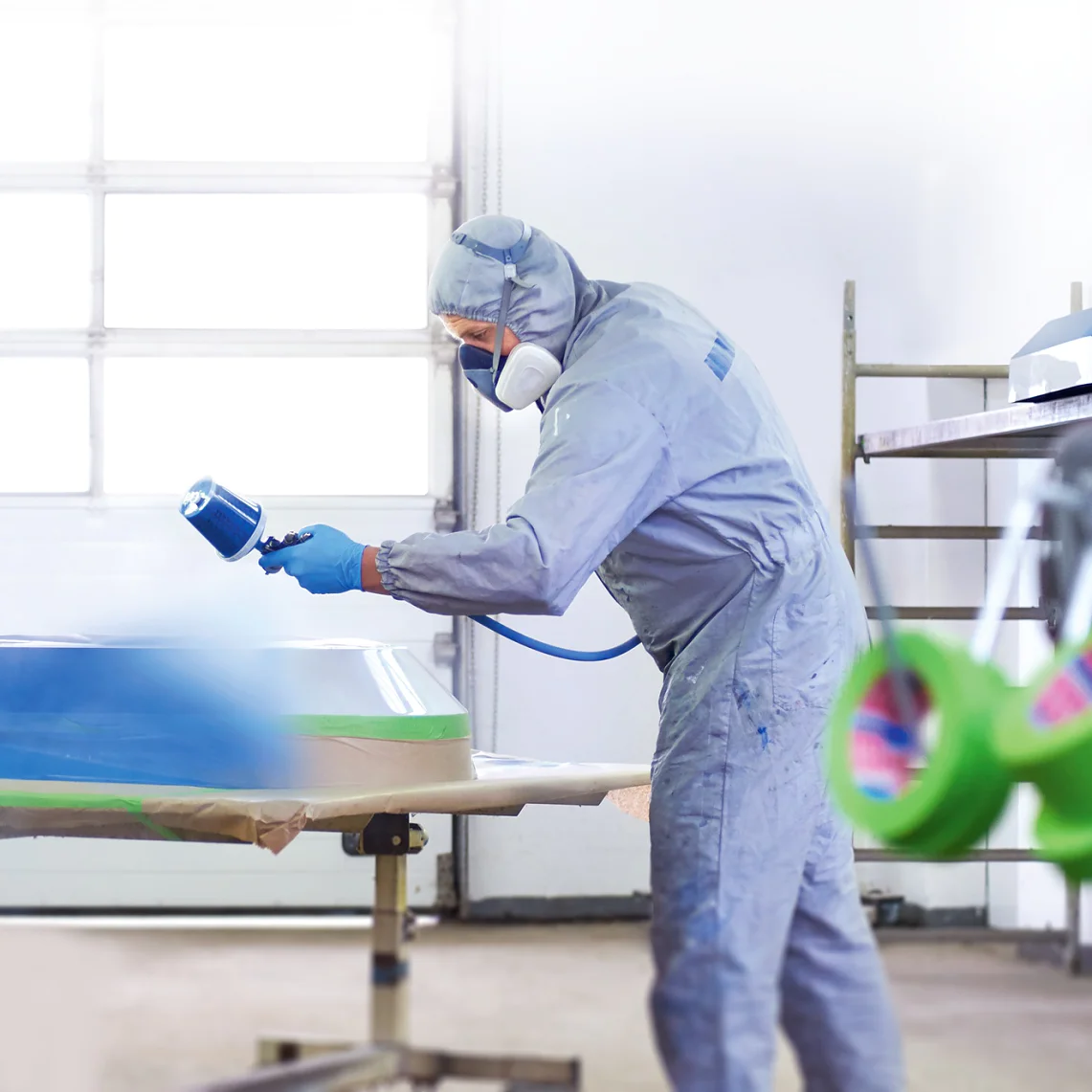 A person in protective clothing and a respirator is spray painting a large object in a workshop. The object rests on a table with tesa tape securing plastic sheeting around the edges. Shelves and equipment are visible in the background. (This text has been generated by AI)