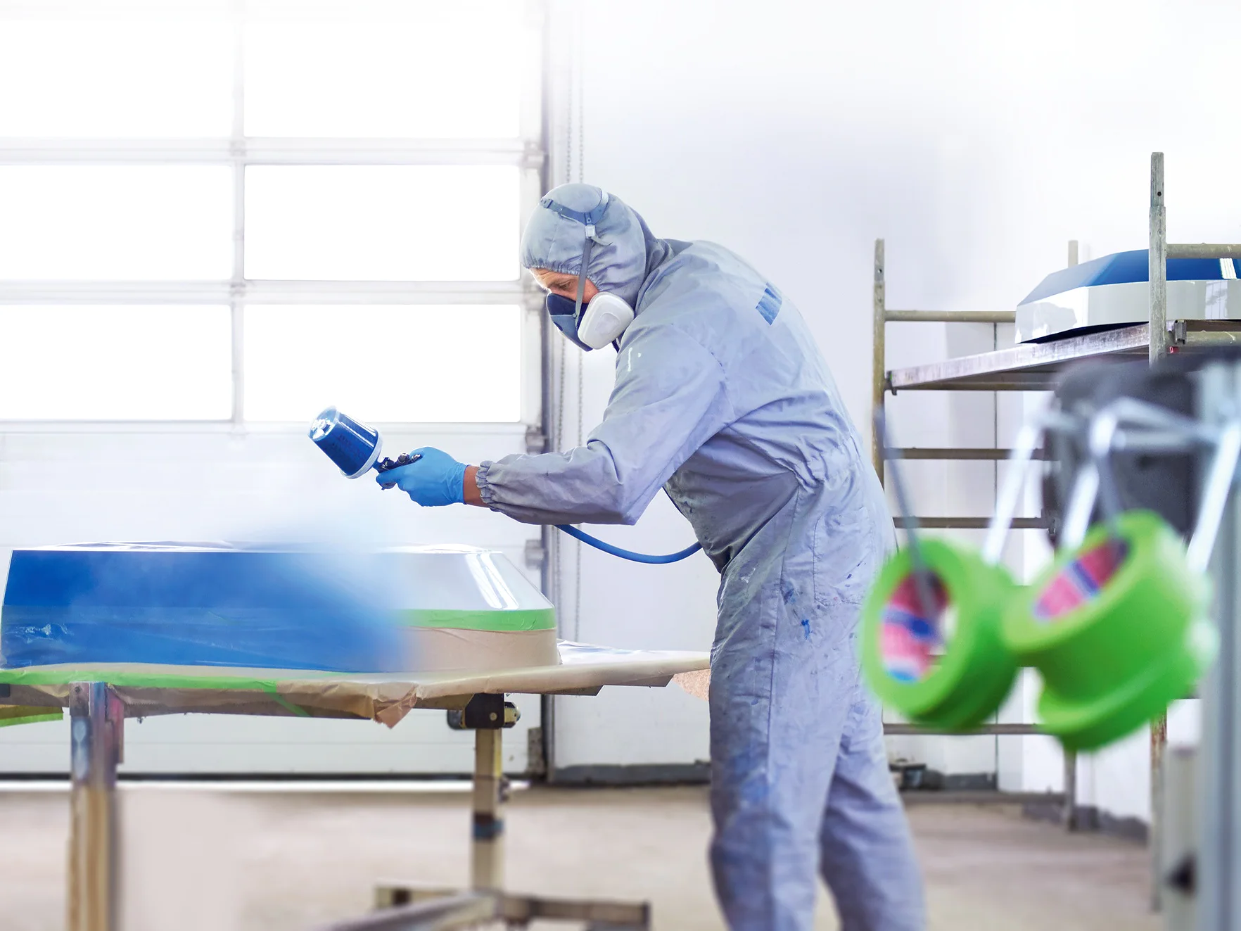 A person in protective clothing and a respirator is spray painting a large object in a workshop. The object rests on a table with tesa tape securing plastic sheeting around the edges. Shelves and equipment are visible in the background. (This text has been generated by AI)