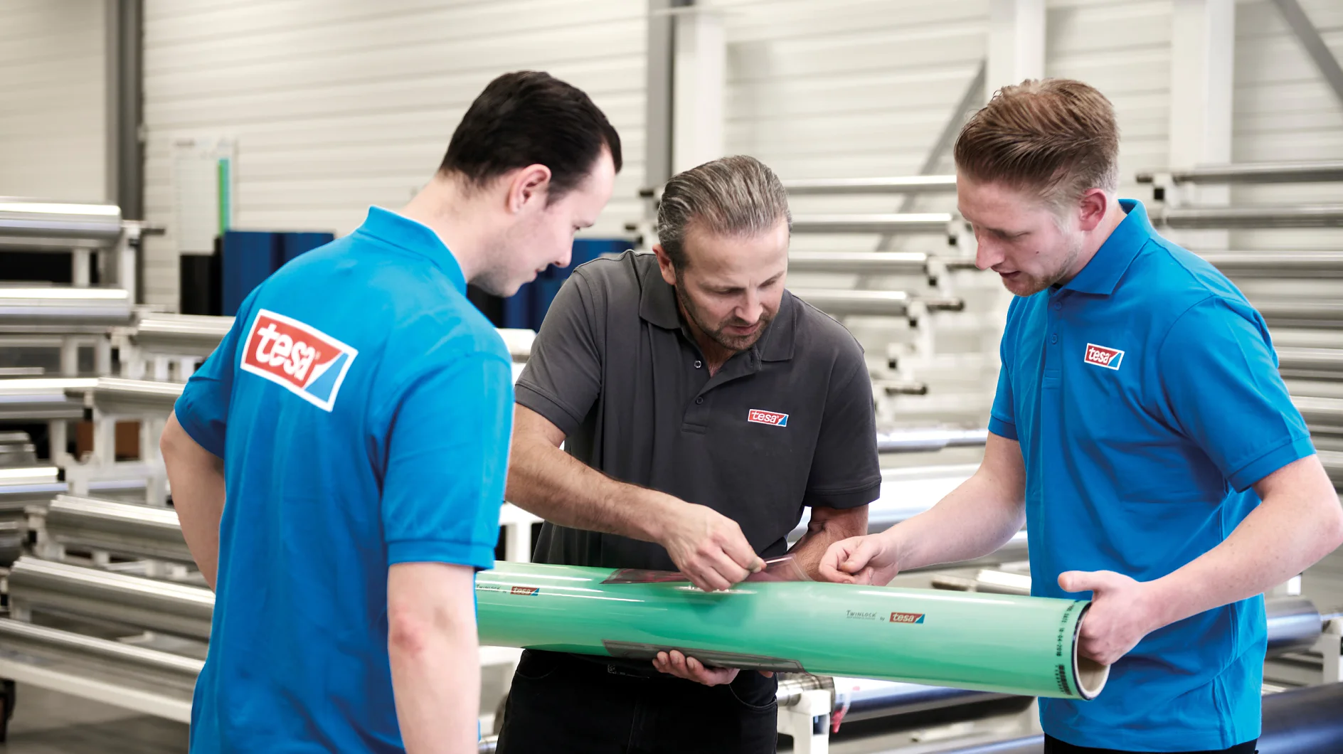 Three men in a factory setting examine a large green cylindrical object. Two men wear blue shirts with red logos, and the third, in the center, wears a gray shirt with the same logo. Metal pipes and machinery are visible in the background. (This text has been generated by AI)