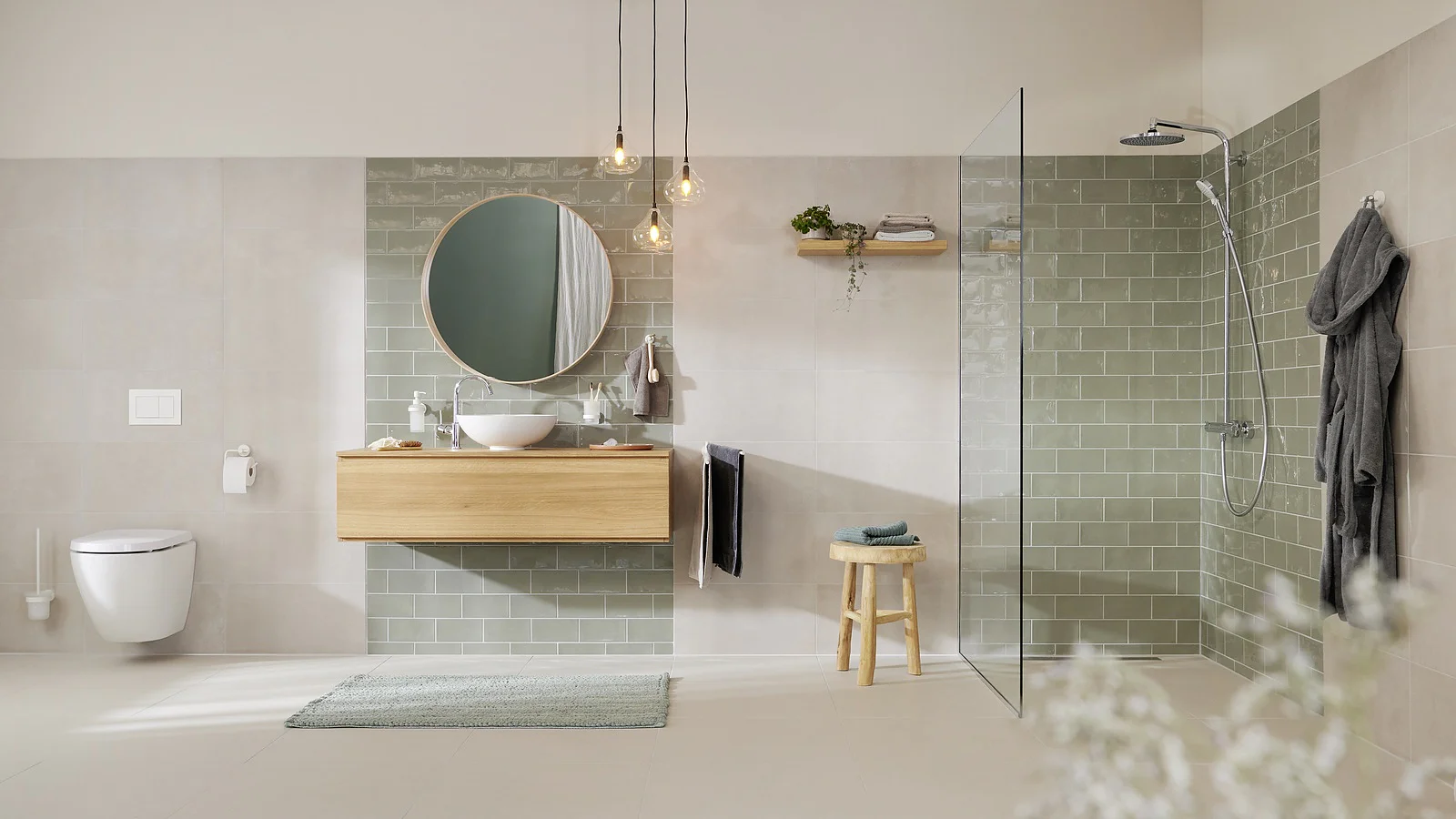 Baño moderno con inodoro montado en la pared, tocador flotante de madera con espejo redondo y ducha con mampara de cristal. Azulejos de color verde claro adornan la pared sobre el lavabo. Lámparas colgantes y un taburete de madera realzan la decoración. Un albornoz sujeto con cinta adhesiva cuelga cerca de la ducha. (This text has been generated by AI)