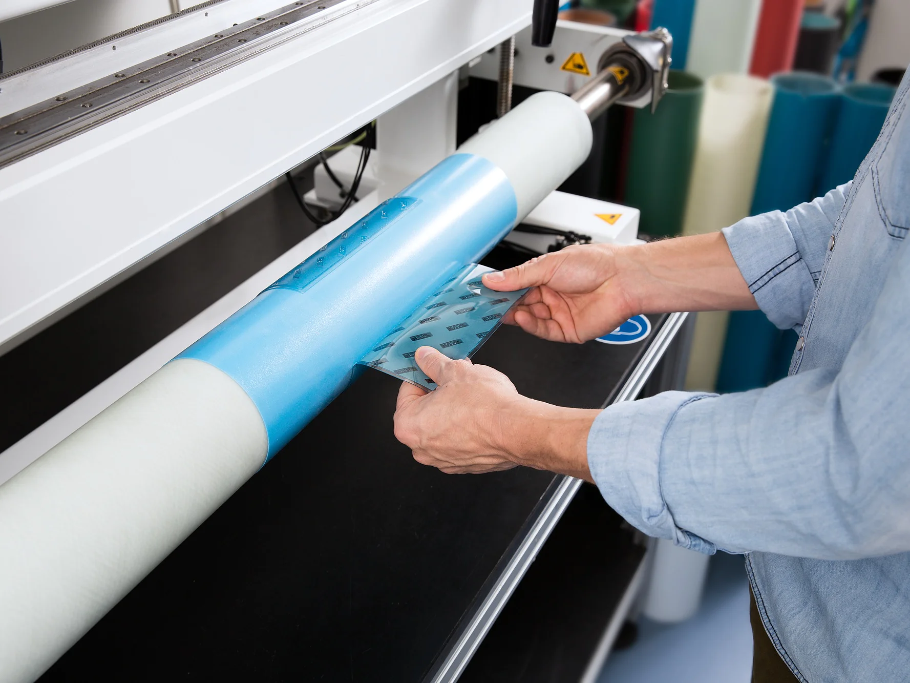flexo-easy-plate-demounting Una persona con una camisa vaquera coloca una película transparente azul sobre una máquina de rollos giratoria en un entorno industrial. La máquina tiene un exterior blanco y metálico con rollos de varios colores en el fondo, incluida una cinta tesa. (This text has been generated by AI)