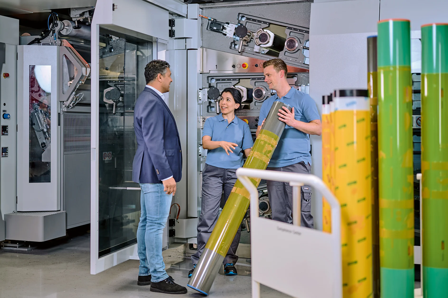 Flexo_Plate-mounting_Consultation_Bobst2023_002d-30777_V1-png_midsize Tres personas se encuentran en una imprenta. Dos trabajadores con uniformes azules manipulan un gran componente cilíndrico de impresión con cinta tesa, mientras un hombre con chaqueta azul y vaqueros observa. Al fondo se ven máquinas y rollos apilados de material impreso. (This text has been generated by AI)