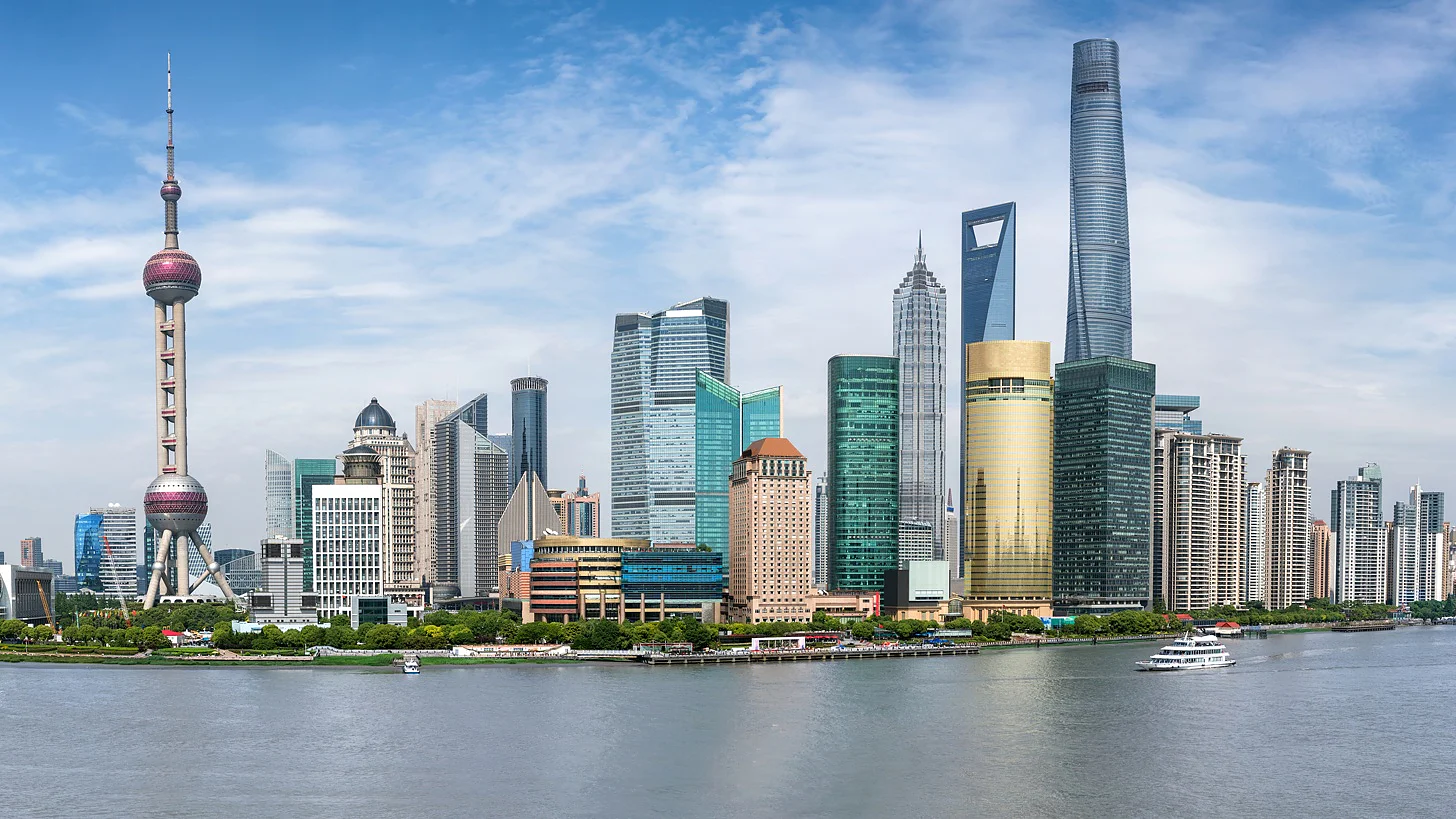 Panoramic view of Shanghais skyline featuring the Oriental Pearl Tower and various skyscrapers along the waterfront, under a clear blue sky. The Huangpu River is in the foreground with boats visible on the water. (This text has been generated by AI)