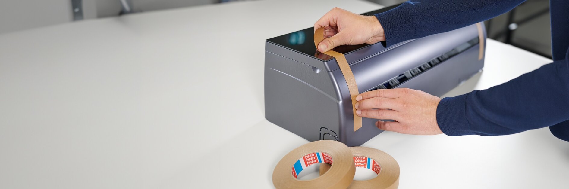 A person is applying a strip of brown tesa tape to the side of a gray machine on a white table. Two rolls of brown tesa tape are placed on the table nearby. (This text has been generated by AI)