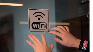 Dos manos colocan una pegatina con el símbolo de Wi-Fi y el texto Wi-Fi sobre una superficie de vidrio con cinta adhesiva Tesa. El fondo está difuminado y muestra parte de un interior. (This text has been generated by AI)