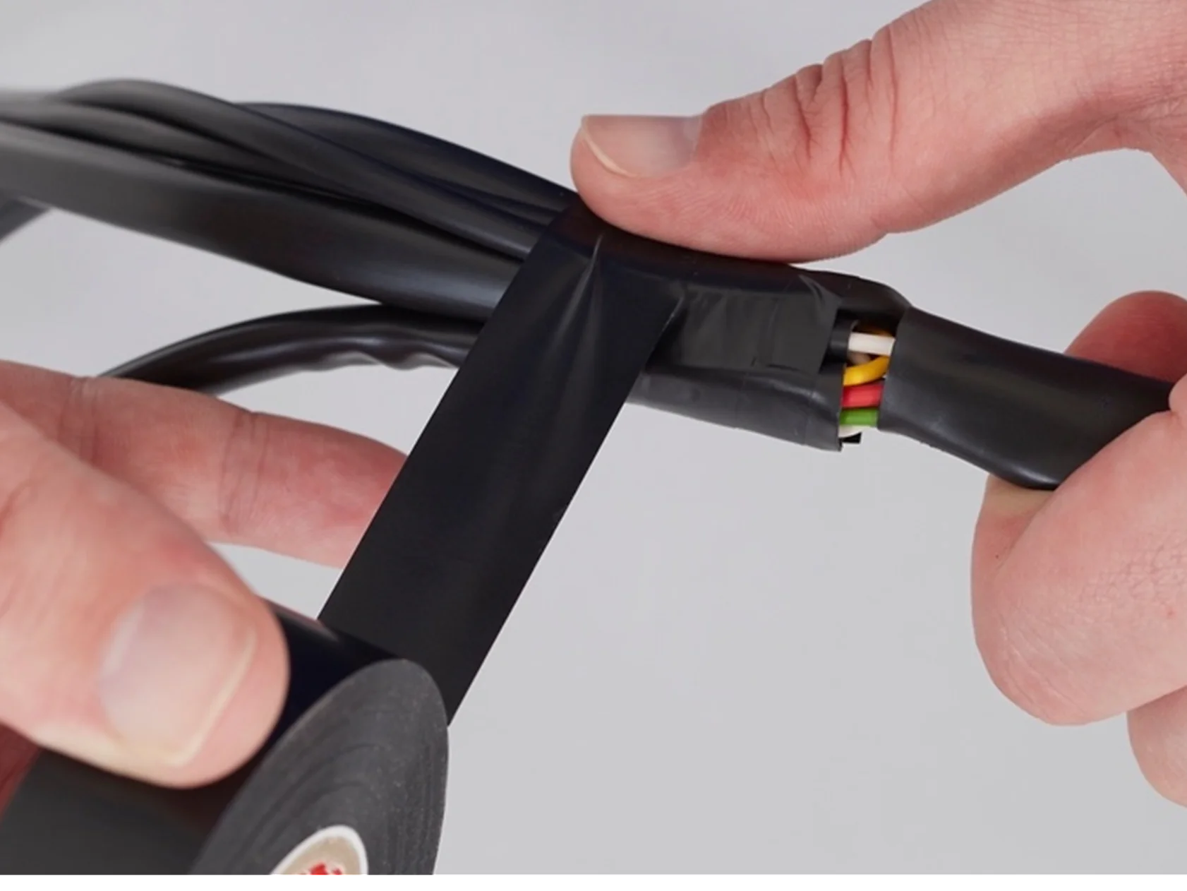 A person wraps black tesa tape around a group of colored wires. The tesa tape is being applied to secure and insulate the wires, which include green, yellow, and red colors. The background is plain white. (This text has been generated by AI)