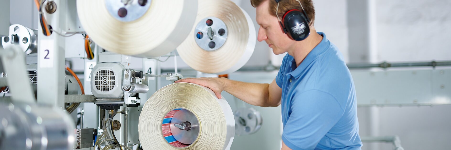 A man wearing a blue shirt and protective earmuffs operates machinery in an industrial setting. He is handling a large spool of tesa tape. There are multiple spools of tesa tape on the machine, and various mechanical components are visible.