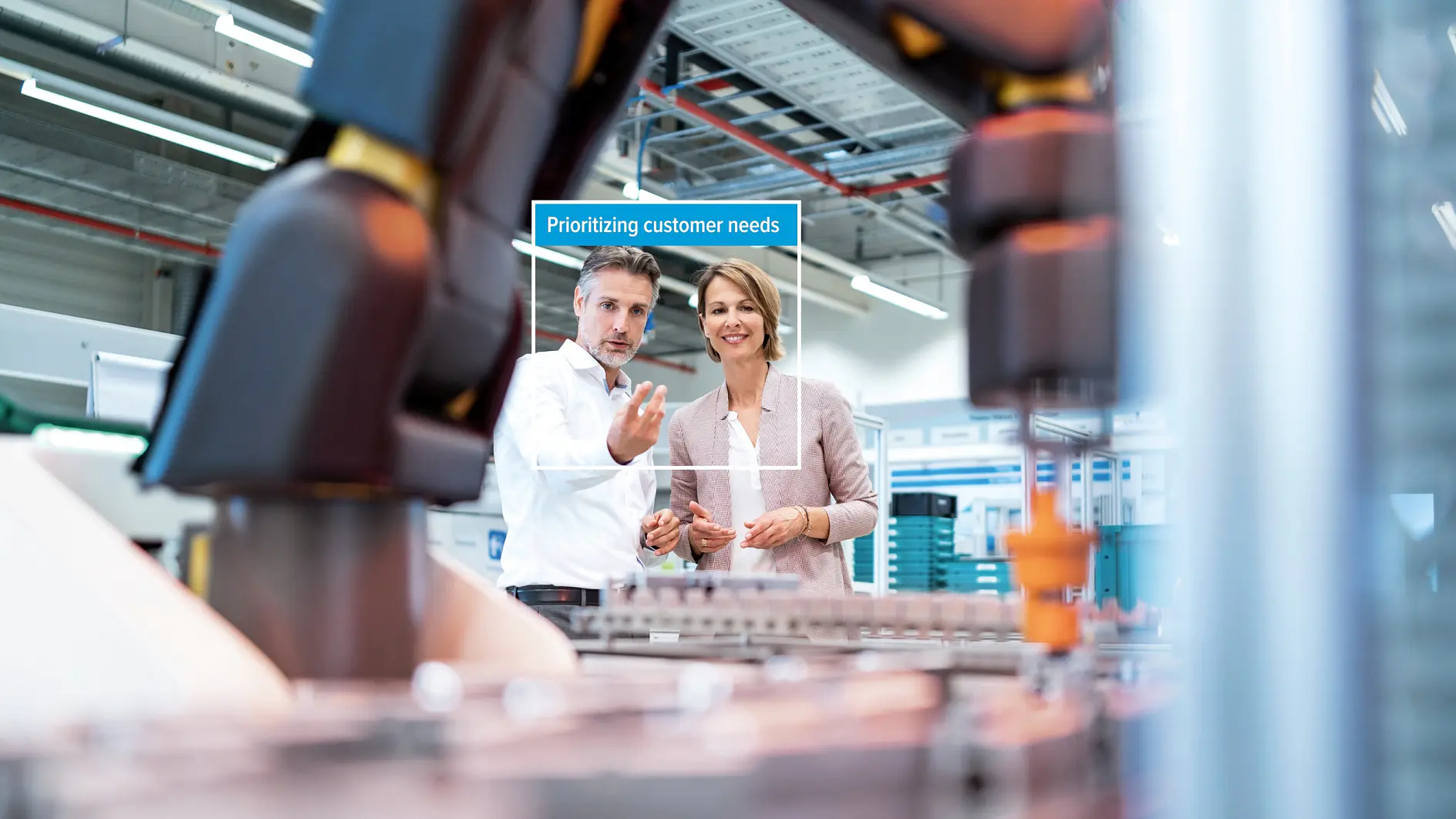 Dos personas de pie en un entorno industrial moderno, cerca de grandes piezas de maquinaria, hablan de la cinta tesa. La pantalla dice: Priorizando las necesidades del cliente. (This text has been generated by AI)