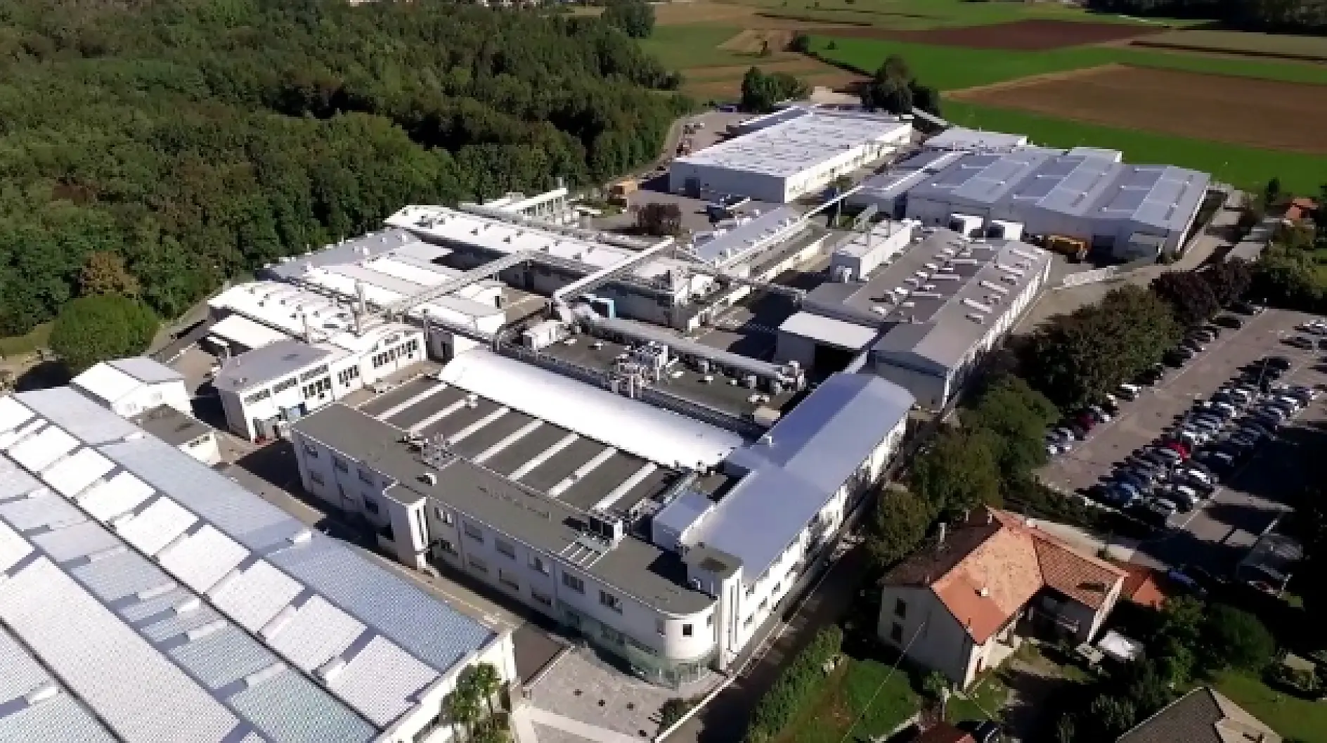 Vista aérea de un gran complejo industrial en medio de campos verdes y árboles, con edificios de tejados blancos y un aparcamiento lleno de coches.