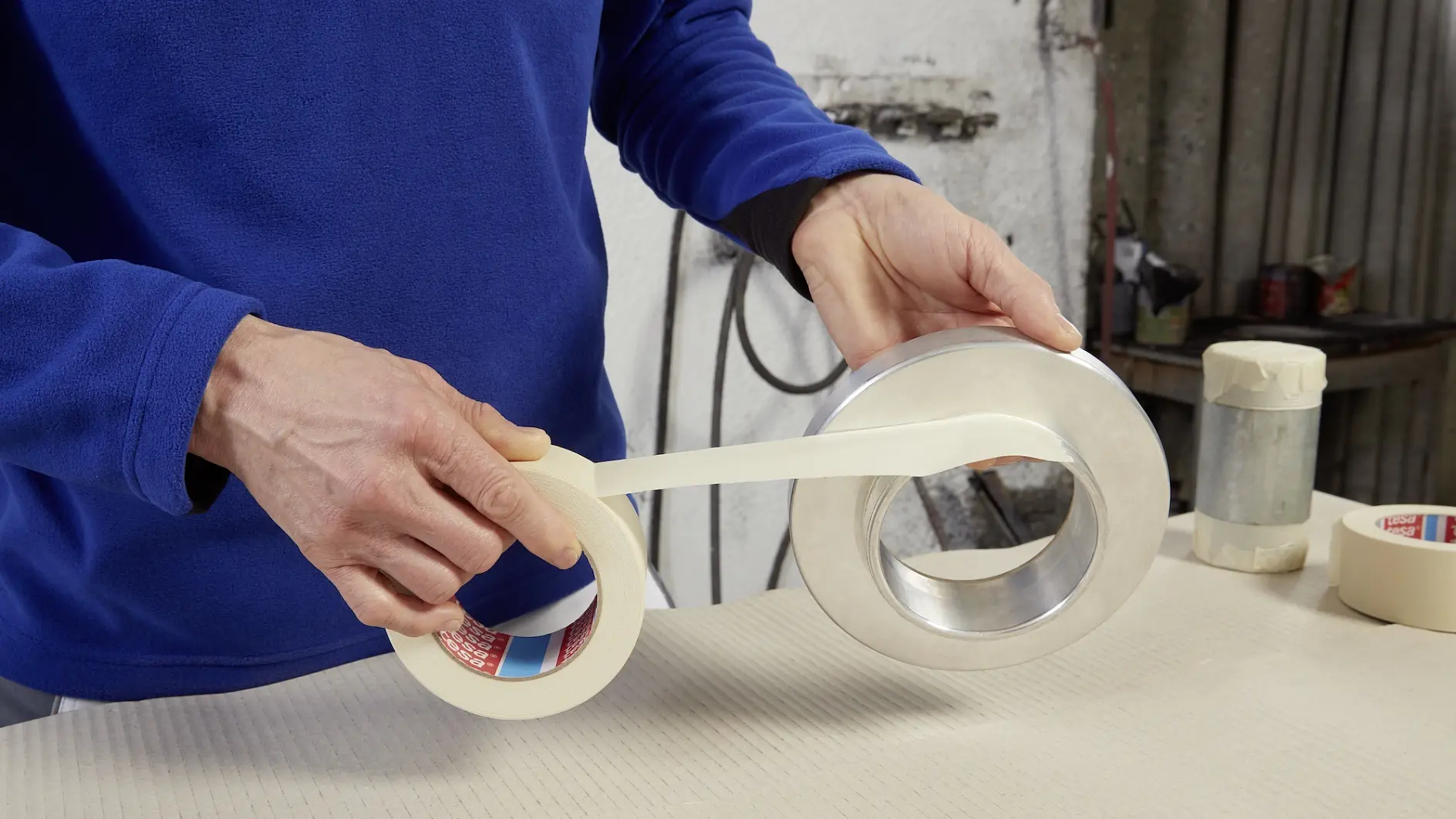 Una persona con jersey azul está aplicando cinta tesa a un objeto metálico circular sobre una superficie beige. Sobre la mesa también hay dos rollos de cinta tesa y un objeto cilíndrico.