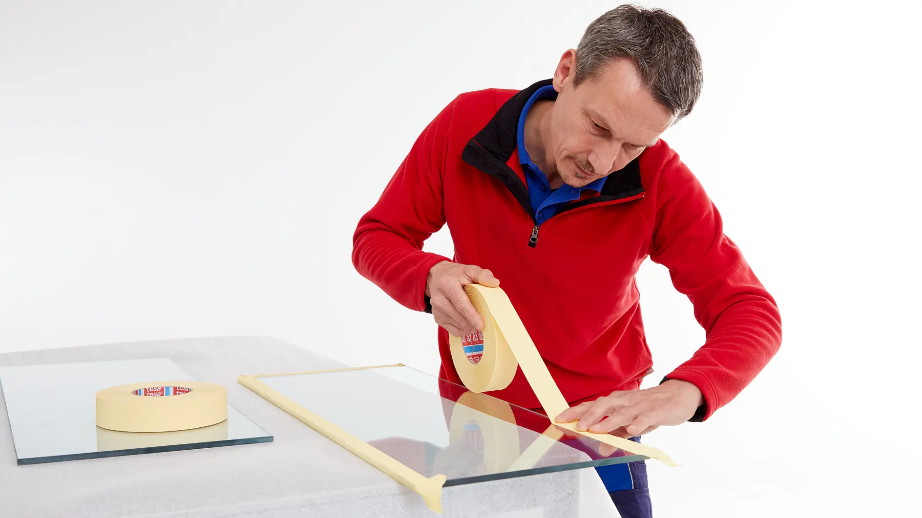Un hombre con chaqueta roja aplica cinta tesa a lo largo del borde de una lámina de cristal sobre una mesa, con otro rollo de cinta tesa y una lámina de cristal cerca.
