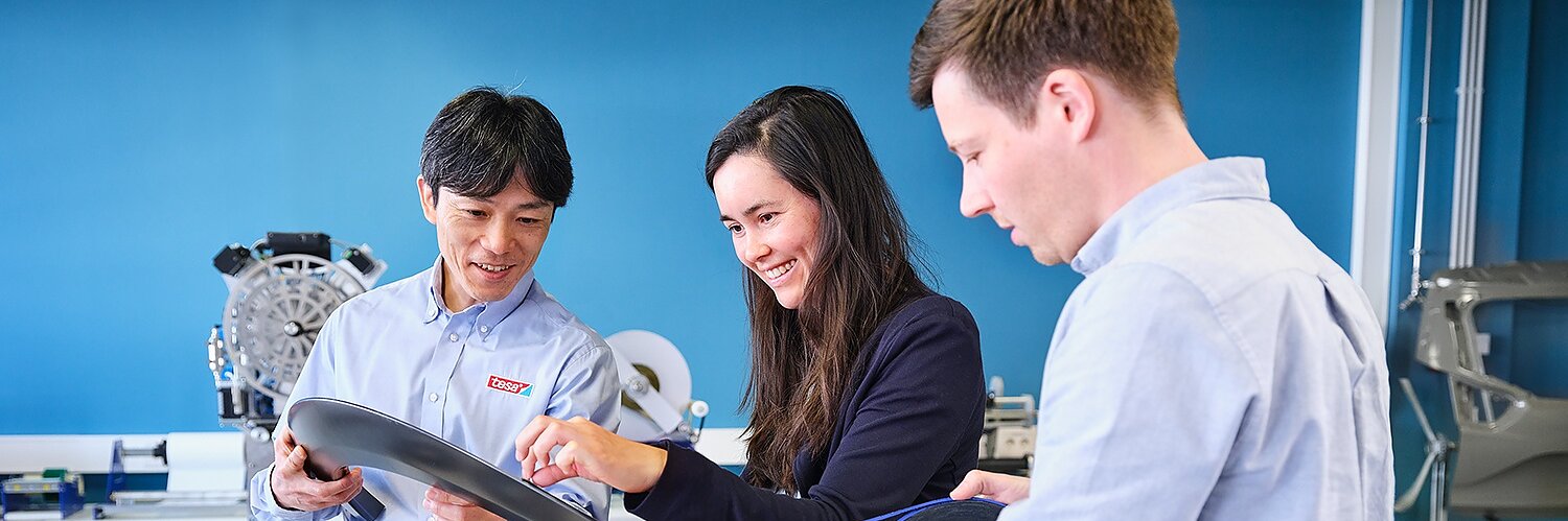 Three people stand at a table examining automotive parts and tesa tape in a bright, modern workspace with blue walls and equipment.