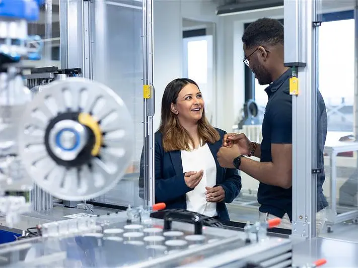 Dos personas se encuentran de pie en un entorno industrial o de laboratorio moderno, conversando. A su alrededor hay maquinaria y equipamiento, entre los que se aprecian rollos de cinta tesa, lo que indica que se trata de un entorno técnico o de fabricación en el que se utiliza la cinta tesa. (This text has been generated by AI)