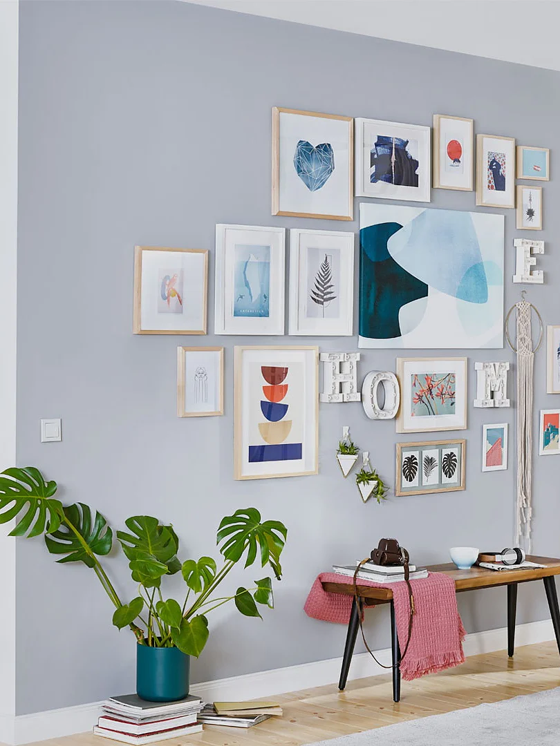 A woman in a pink shirt is arranging frames on a grey and white wall, filled with various artwork and photos, using tesa tape. The room has a modern design with a small kitchen on the left, light wood flooring, and green plants for decoration. (This text has been generated by AI)