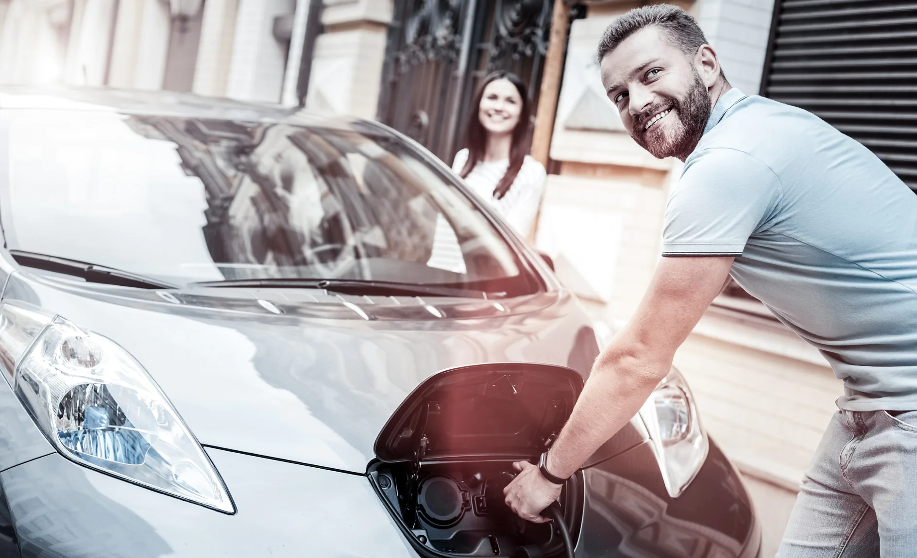 E-Mobility Key Visual Easy and ecological transportation. Selective focus on a happy millennial guy grinning broadly while holding a charging nozzle and enjoying the process of his eco car supplying energy outdoors.