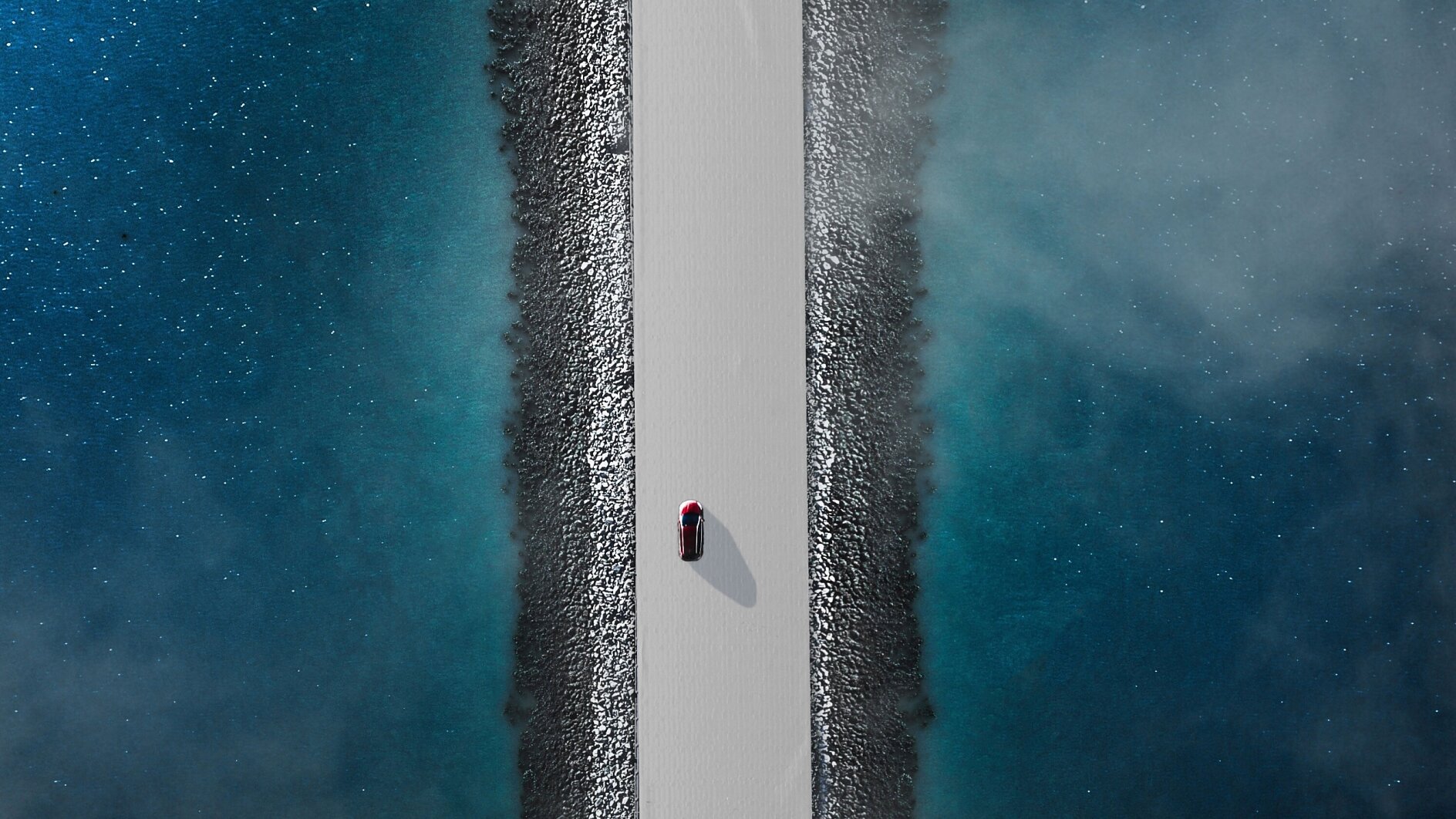 Aerial view of a red car driving on a narrow, straight road flanked by blue water on both sides. The road is elevated, with rocky edges lining the sides. The water appears calm with a slight mist or fog hovering above it. (This text has been generated by AI)