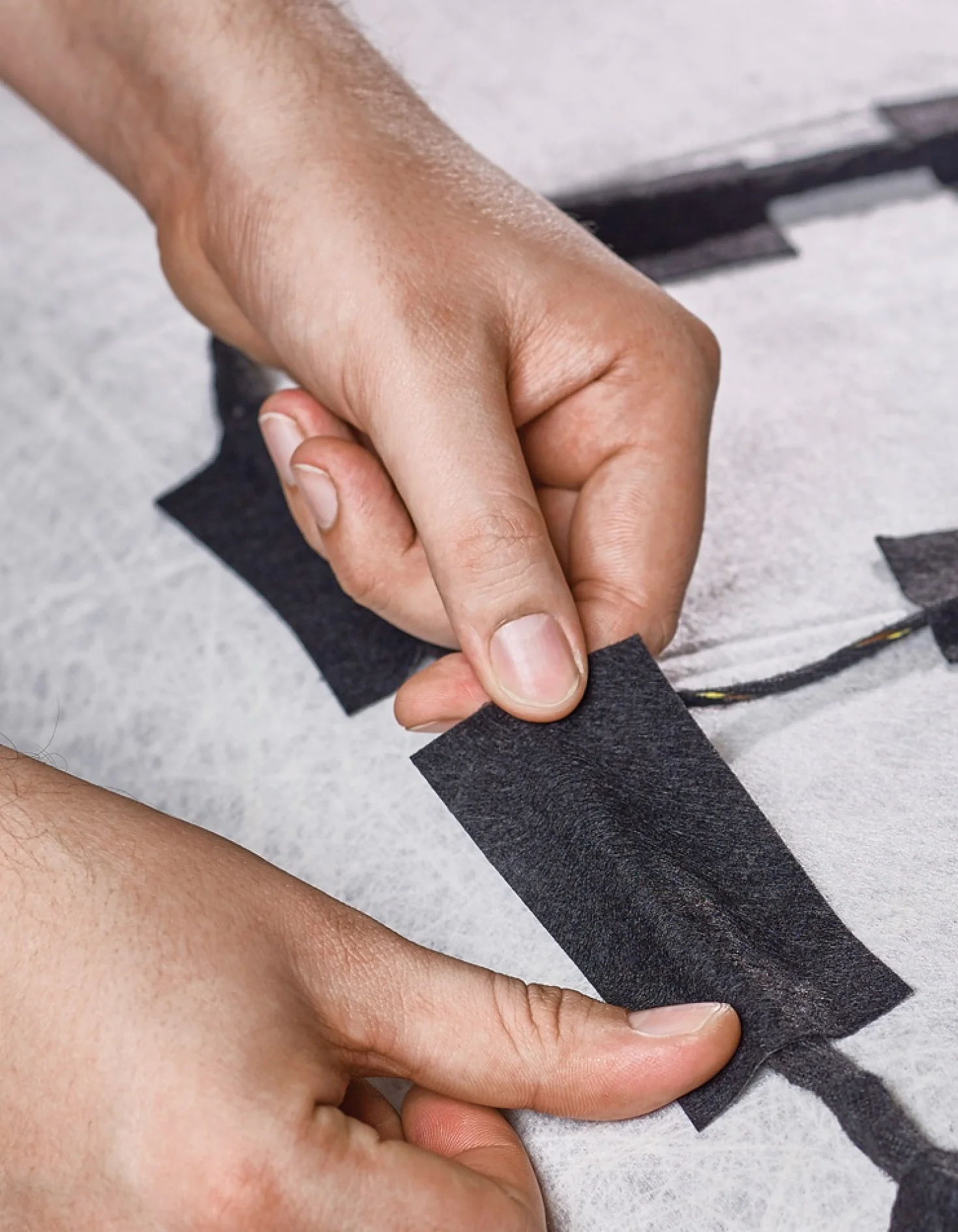 Primer plano de dos manos aplicando un trozo rectangular de cinta tesa negra sobre una superficie blanca texturizada, con otras cintas tesa negras cerca. (This text has been generated by AI)