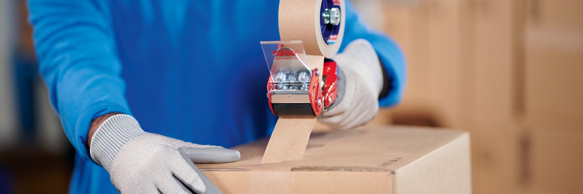 A person wearing a blue jacket and gray gloves is sealing a cardboard box with brown packing tesa tape using a red tesa tape dispenser. Other cardboard boxes are visible in the background.