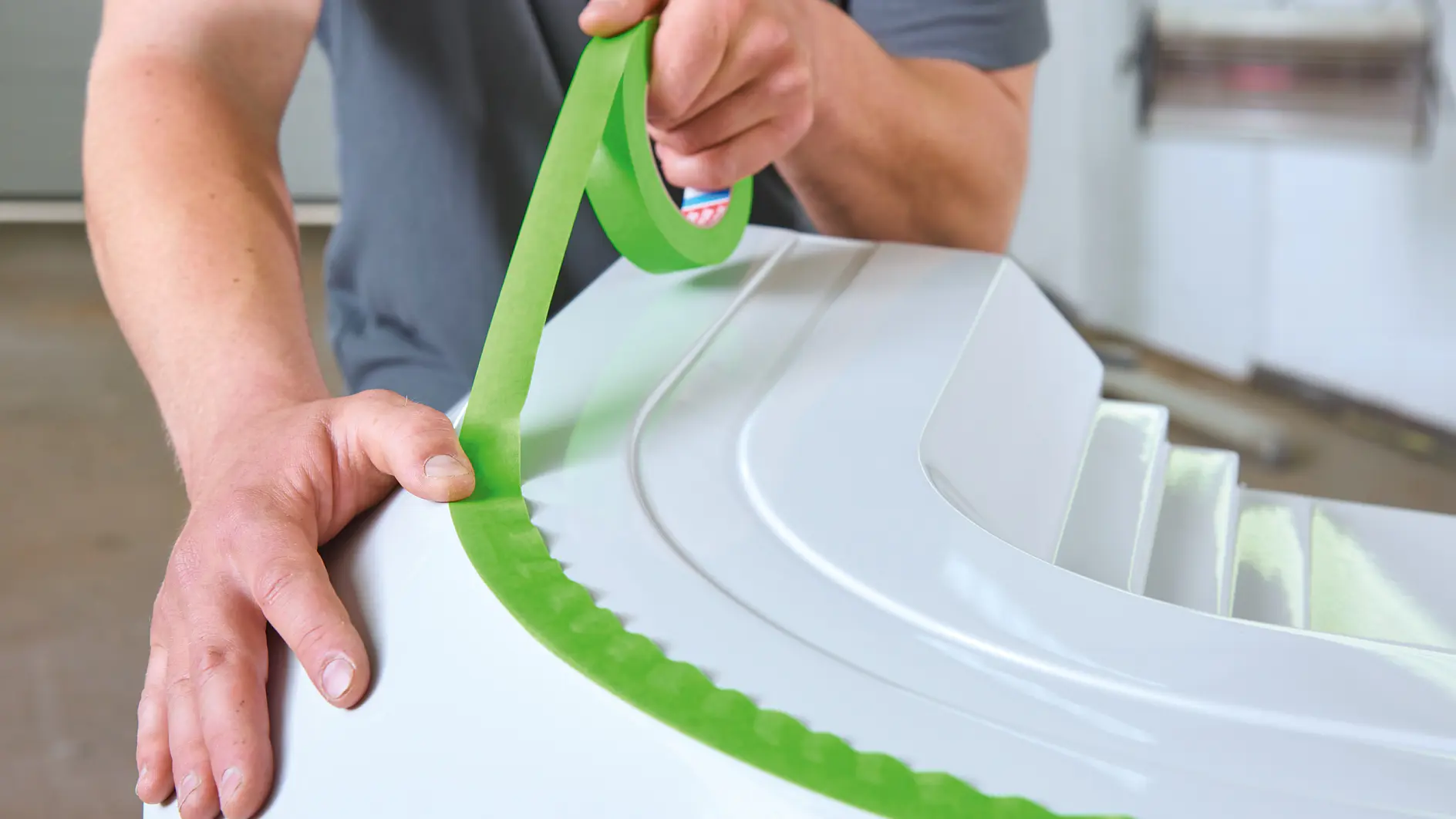 Una persona está aplicando cinta tesa verde al borde de una superficie blanca, utilizando ambas manos para alinearla cuidadosamente. La persona lleva puesta una camiseta gris y está completamente concentrada en el proceso de aplicación de la cinta tesa.