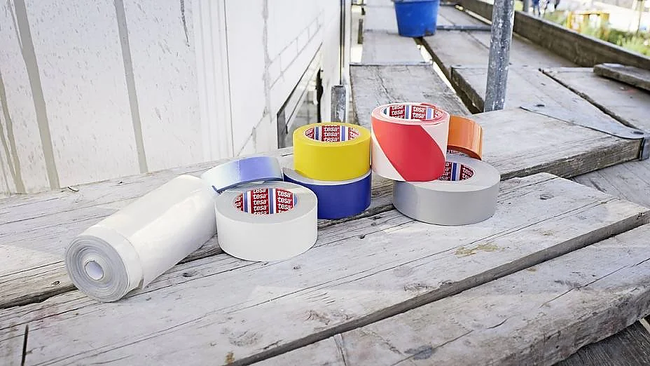 Erinevad värvilise tesa teibi rullid, sealhulgas sinine, kollane, punane, hõbedane ja valge, koos valge plastlehe rulliga asetatakse ehitusplatsil puitlaudadele. Taustal on näha sinine kopp ja tellingud. (This text has been generated by AI)