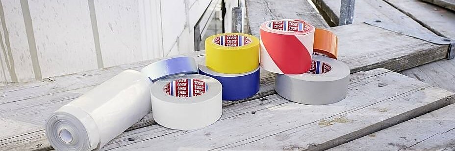 Erinevad värvilise tesa teibi rullid, sealhulgas sinine, kollane, punane, hõbedane ja valge, koos valge plastlehe rulliga asetatakse ehitusplatsil puitlaudadele. Taustal on näha sinine kopp ja tellingud. (This text has been generated by AI)