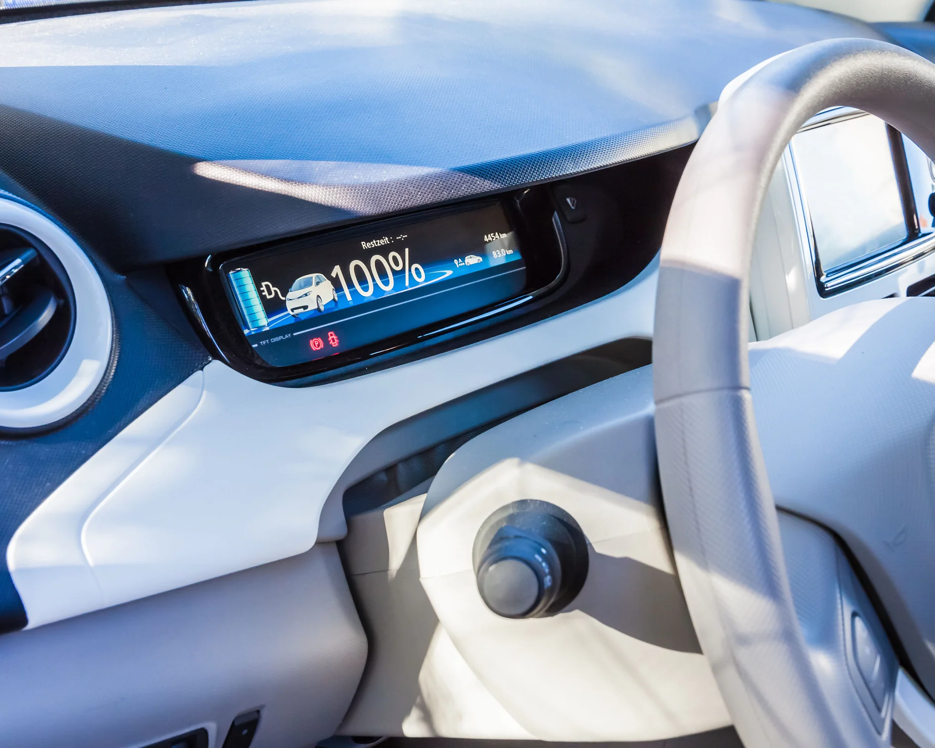 auto_juhikabiin-1 Close-up of a car dashboard featuring a digital screen showing a fully charged electric vehicle battery at 100%. The steering wheel and a round air vent are visible. The interior is light-colored. (This text has been generated by AI)