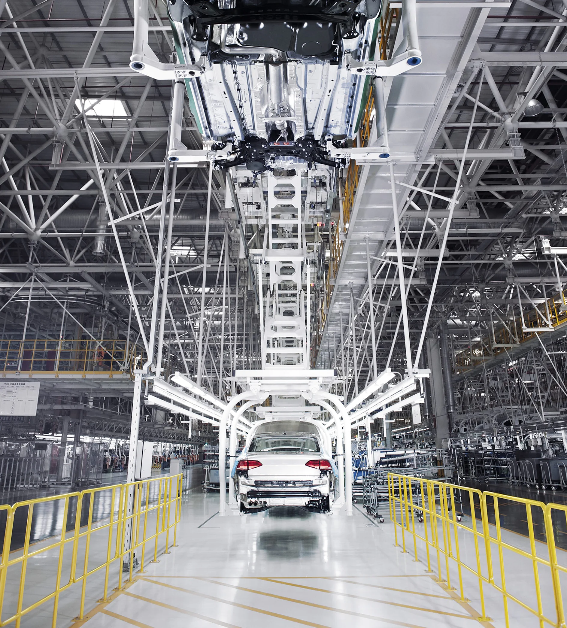 autotehas A white car is on an assembly line in an industrial factory. The overhead structure supports various machinery components. The surrounding area is spacious, with yellow railings along the pathway on each side. Each piece of equipment that requires fastening or securing uses tesa tape for optimal performance and reliability throughout the manufacturing process. (This text has been generated by AI)