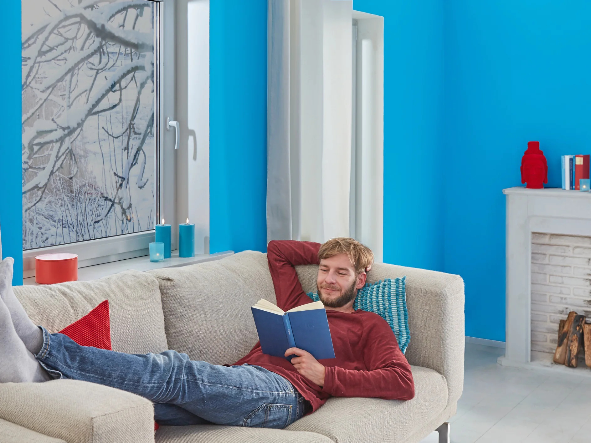 A man is relaxing on a beige couch, reading a book in a room with bright blue walls. He wears a red sweater and jeans, with his legs resting on the couch. In the background, theres a white fireplace and a window showcasing snowy branches. (This text has been generated by AI)