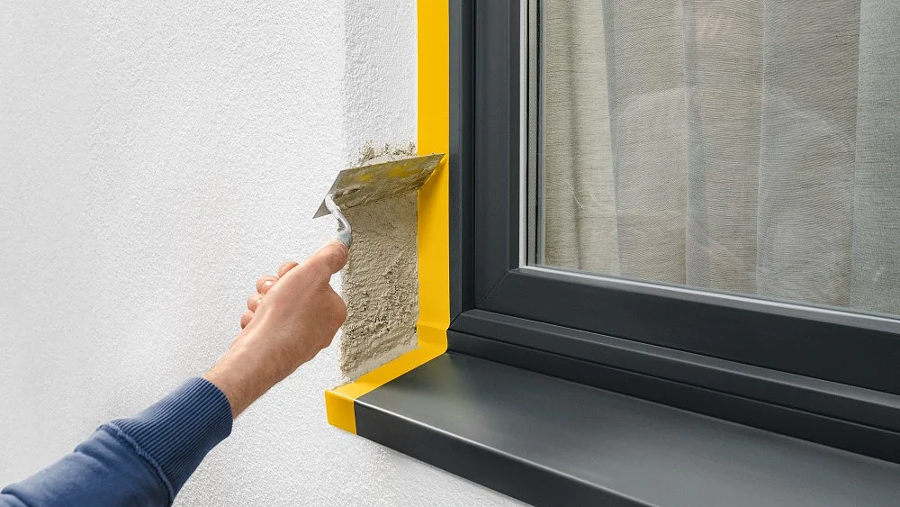 A person applies stucco with a trowel to a wall next to a window with a black frame and sill. The window has yellow tesa tape along the edges, and a reflection of the persons arm is visible in the glass. (This text has been generated by AI)