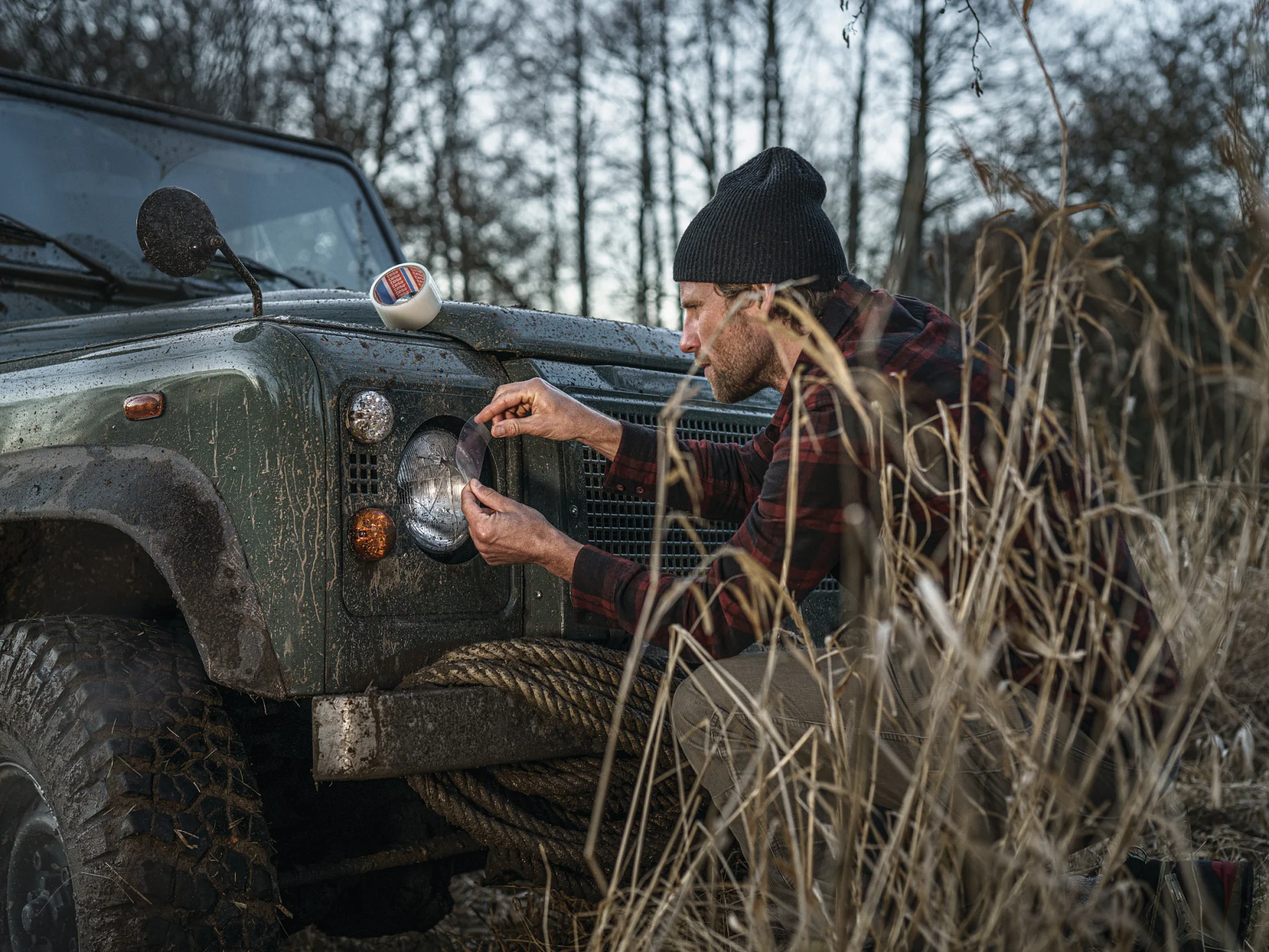 Nokamütsi ja ruudulist särki kandev mees põlvitab maastikusõiduki kõrval ja vahetab esitule pirni. Sõiduk on porine ning ümbrus on võsastunud kõrge rohu ja puudega, mis viitab maa- või põlisloodusele. (This text has been generated by AI)