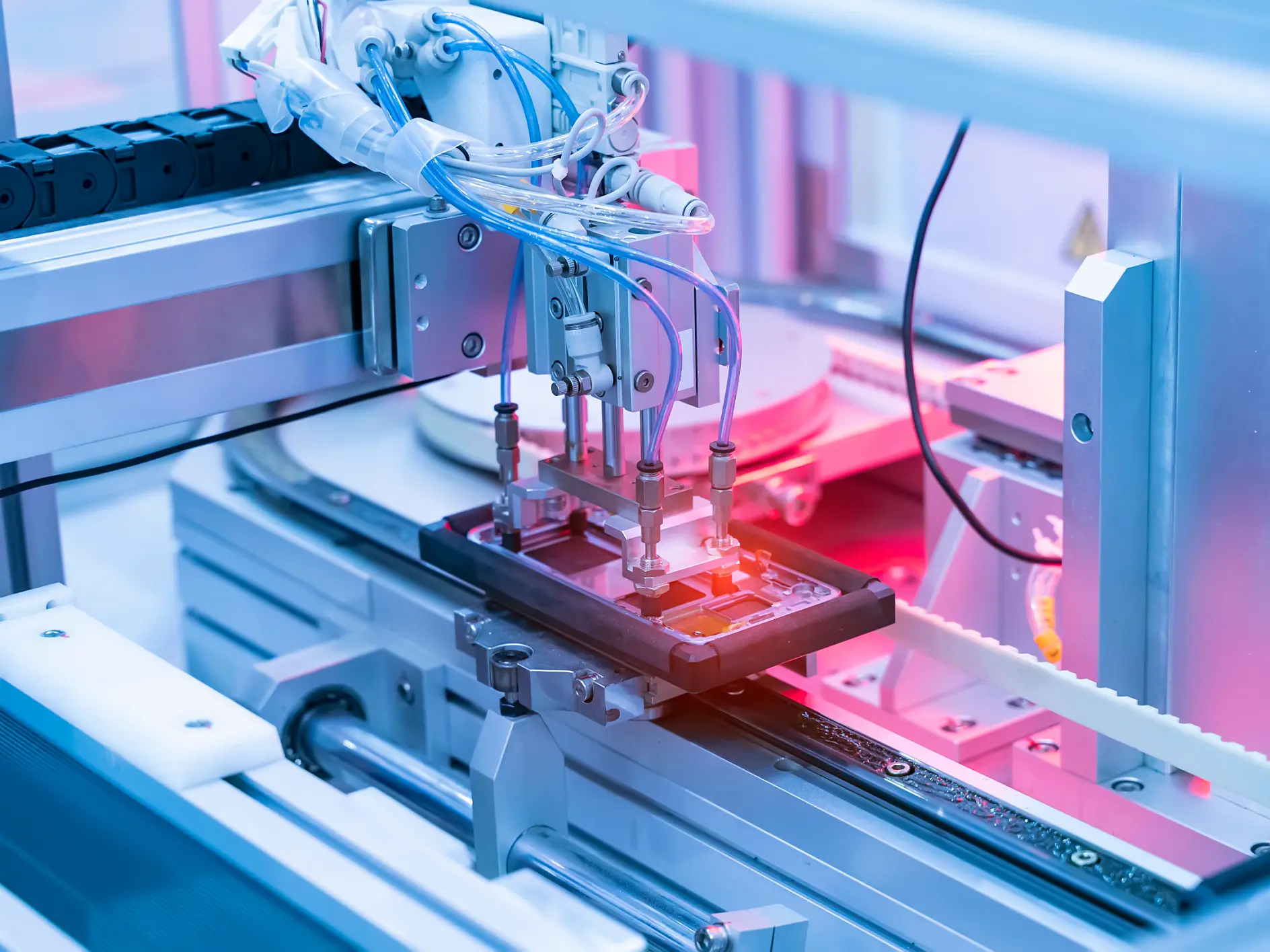A robotic arm operates on an electronic device in a manufacturing facility. The setting is highlighted with pink and blue lights, showcasing precision machinery used in automated production processes. Various cables, mechanical parts, and rolls of tesa tape are visible.