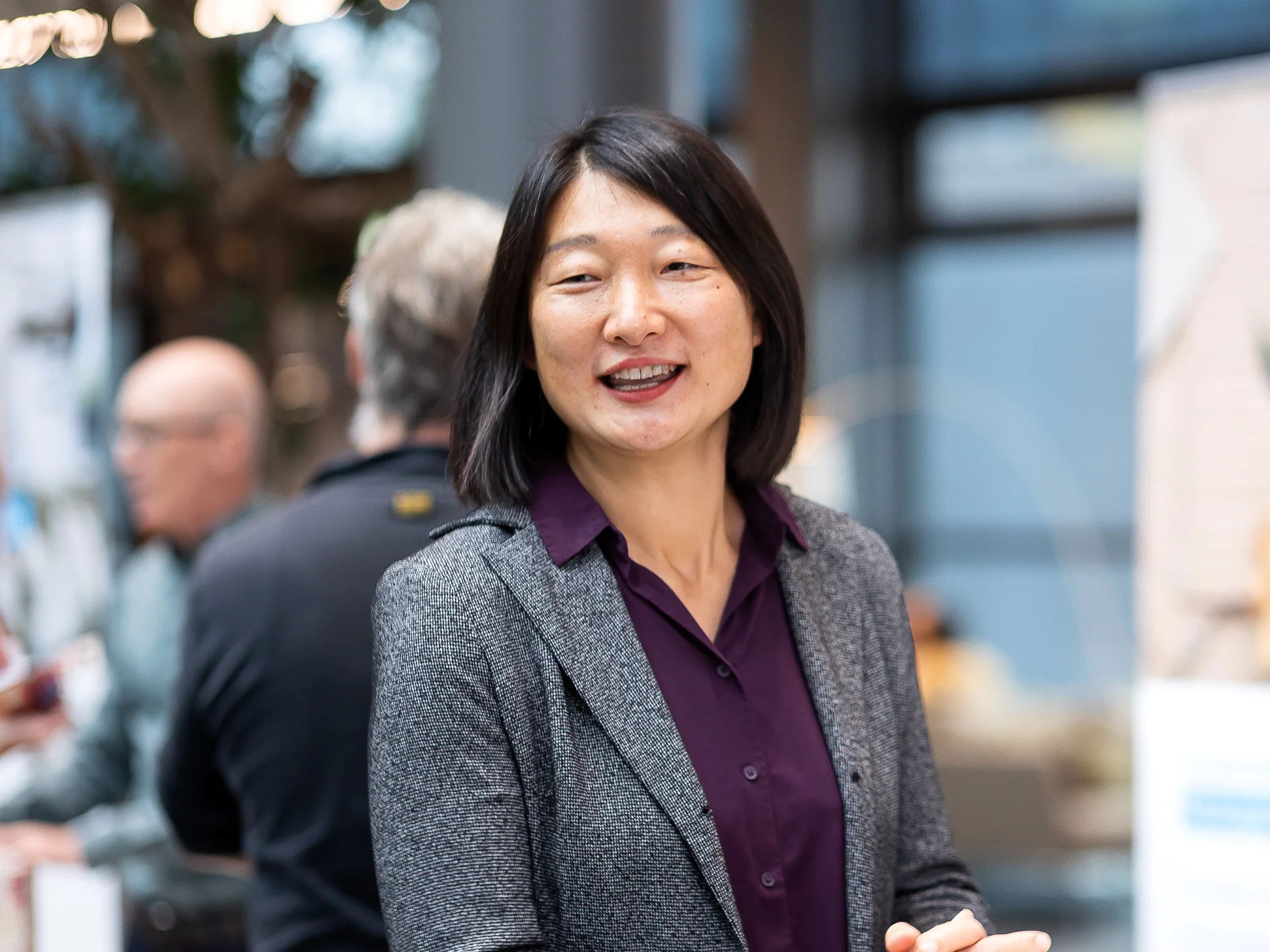 A woman with shoulder-length black hair is smiling, wearing a gray blazer and purple blouse. She is standing indoors, with people and blurred background elements like posters and a window behind her. Please provide the text that contains references to tapes so I can help you make the necessary modifications by using tesa tape. (This text has been generated by AI)