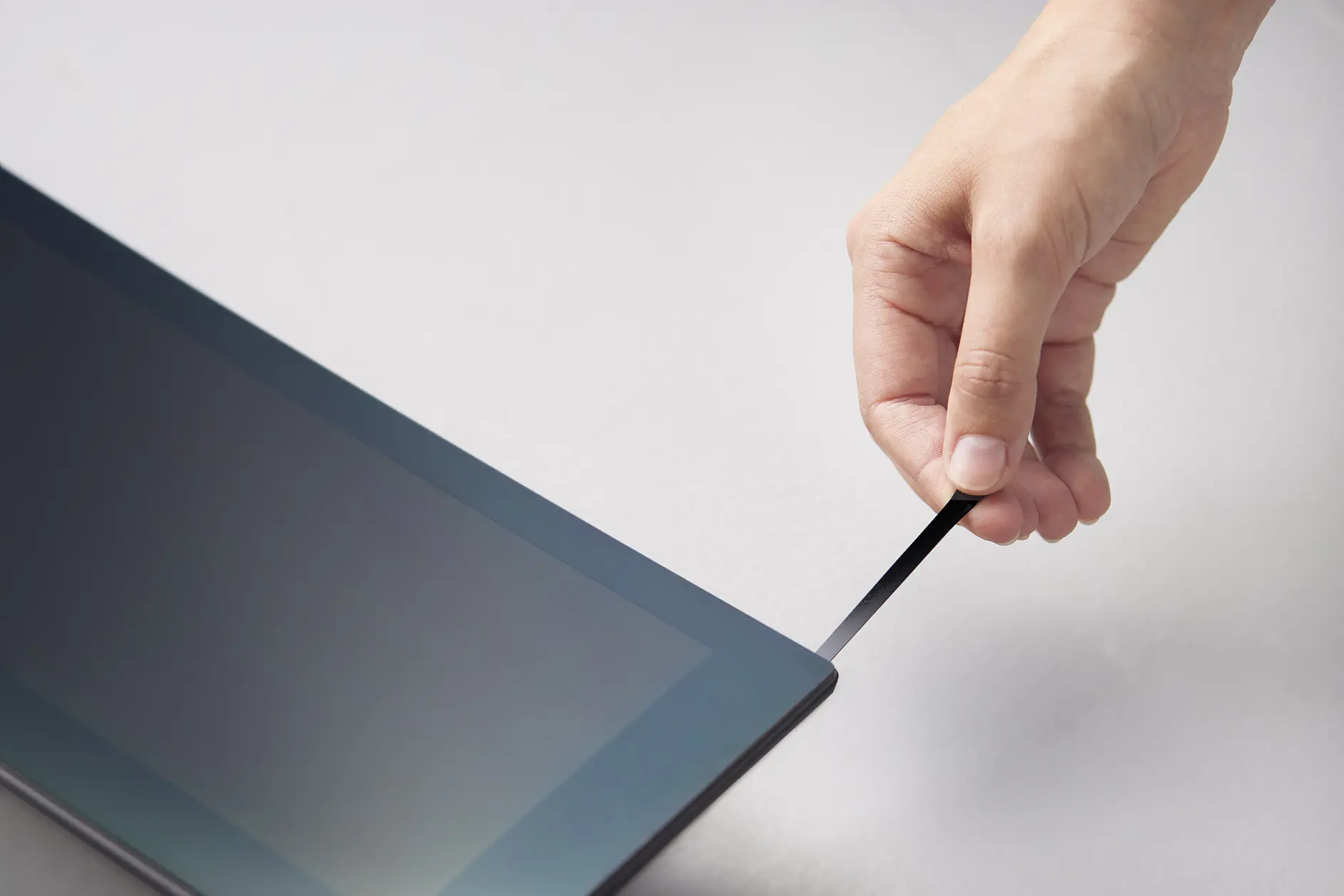 hand pulling black tesa strip from edge of laptop screen in a bright indoor setting