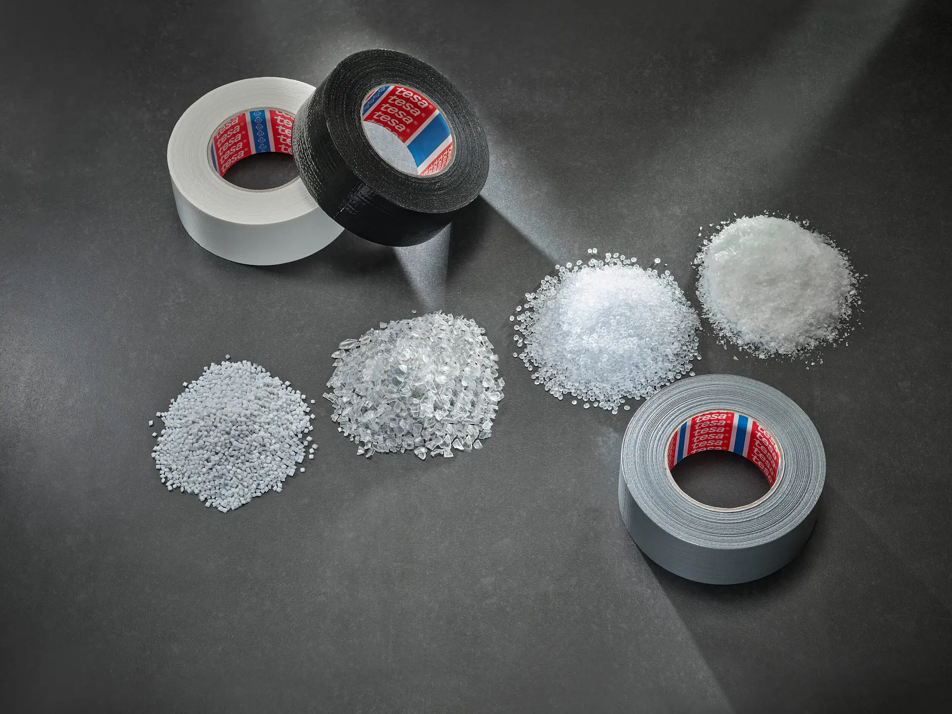 three rolls of tesa adhesive tape in white, black, and gray placed on a dark surface with corresponding piles of white granular material nearby