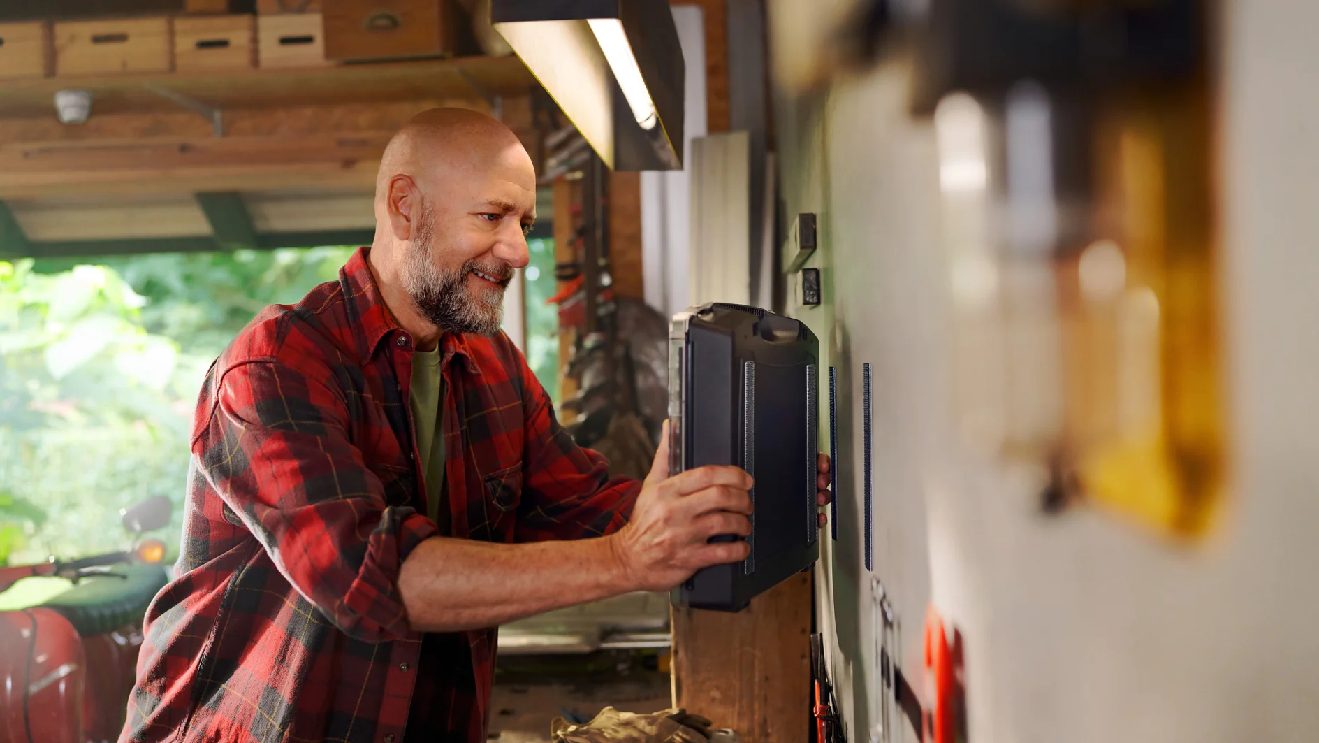 A bald man with a beard, wearing a red plaid shirt, is placing a toolbox on a shelf in his workshop. He smiles as he organizes tools around him. The workshop is brightly lit with various tools and equipment visible in the background, along with tesa tape prominently displayed among them. (This text has been generated by AI)